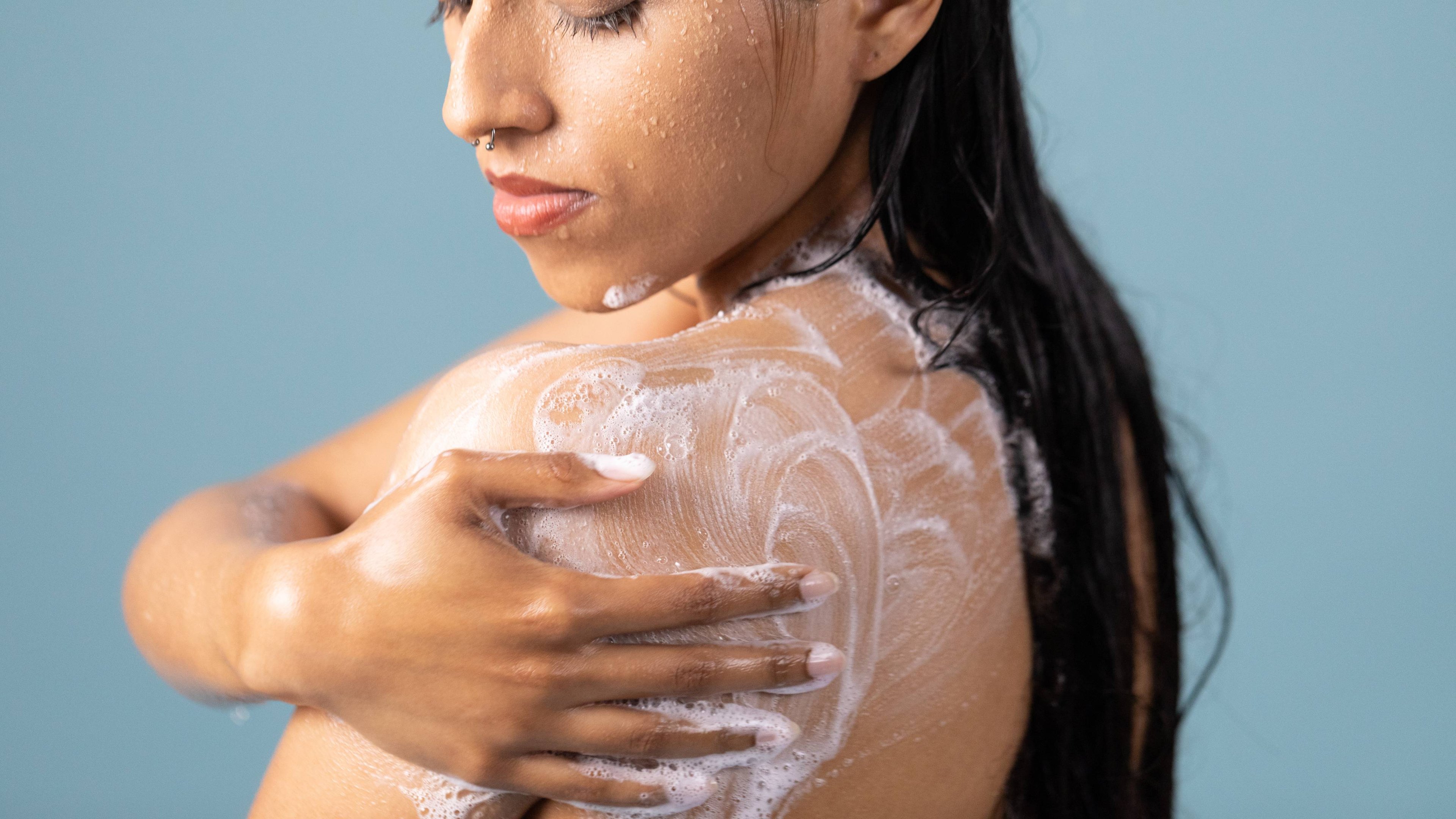 The model is on a pale blue background stood under the shower lathering up their shoulder with shower gel.