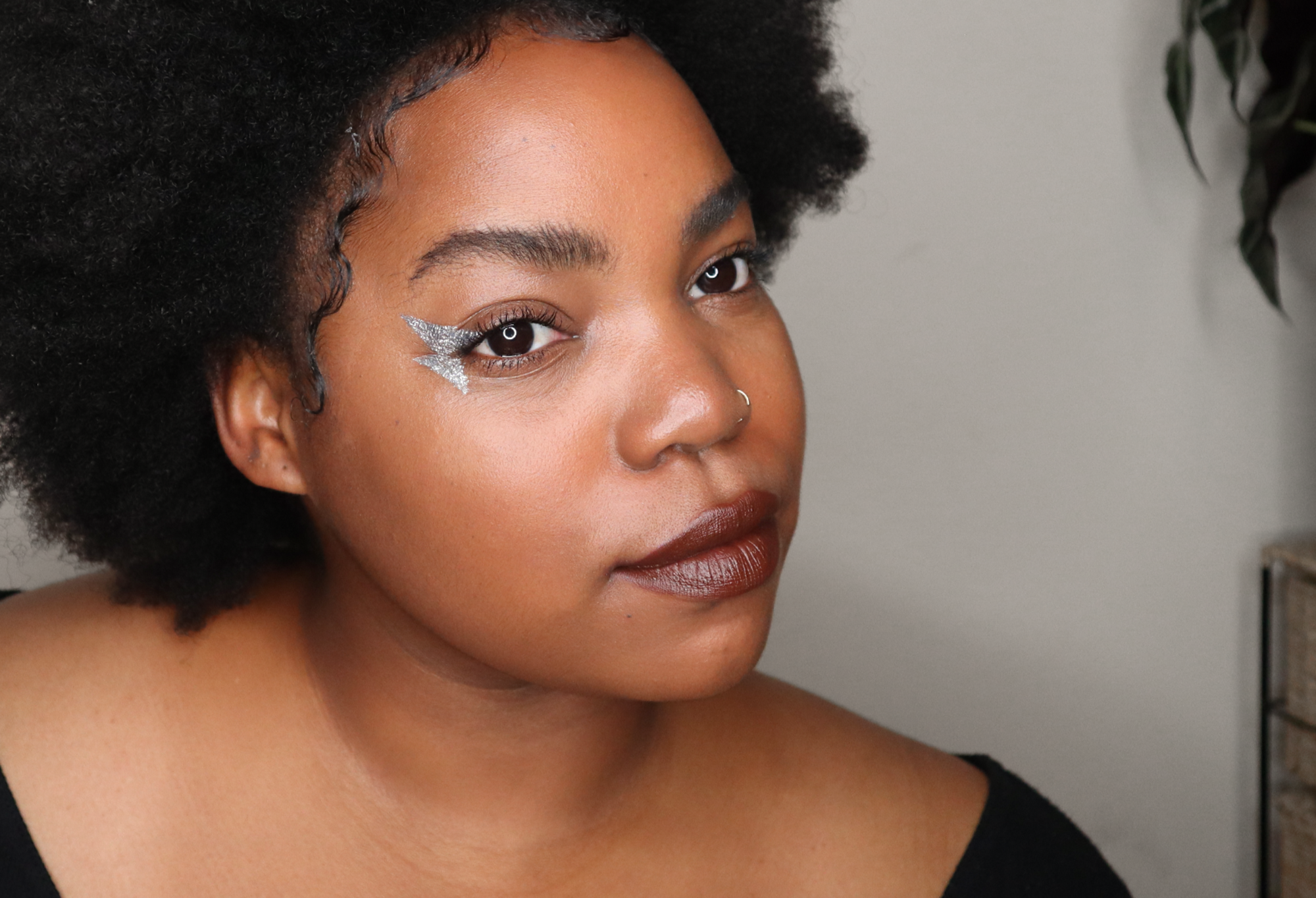 A person with Afro hair and a nose ring looks thoughtful, with a silver lightning bolt painted in the corner of their eye.