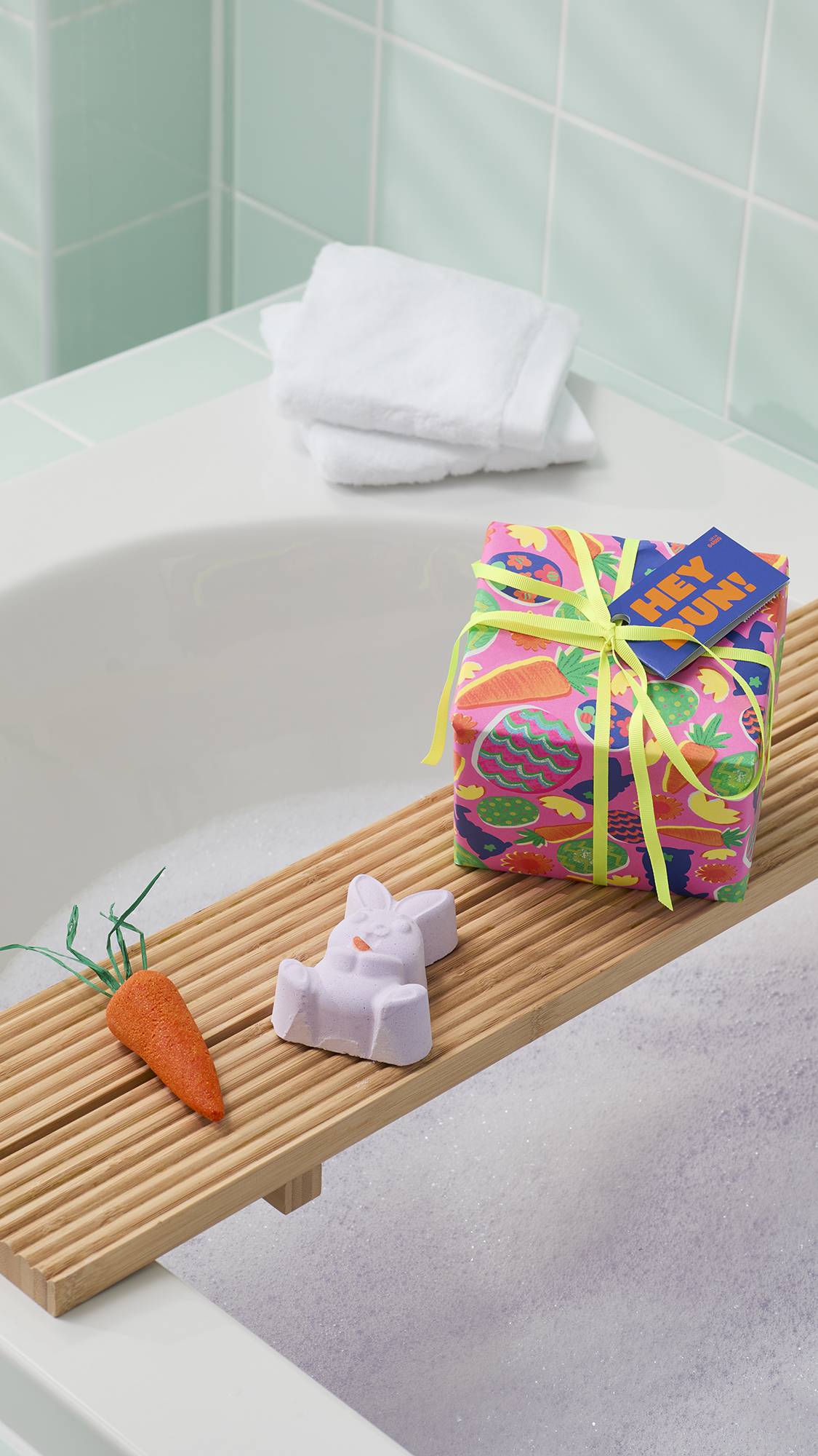 The image shows the Lush "Hey Bun!" gift set on a wooden bath tray over a bubbly tub. The set includes a pink gift box with a "HEY BUN!" tag, a bunny bath bomb, and a carrot-shaped bubble bar. White towels rest on the corner of the bath behind it.