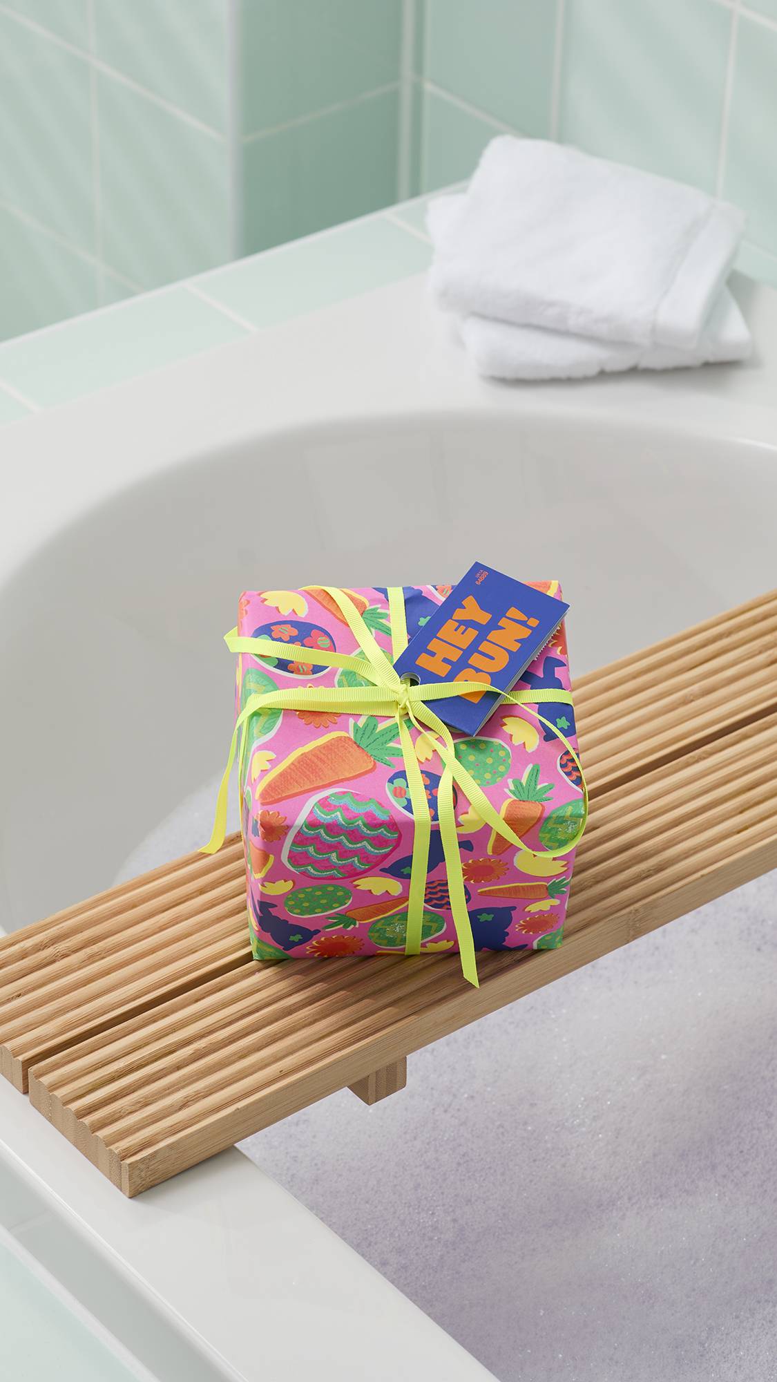 The image shows the Lush "Hey Bun!" gift set on a wooden bath tray over a bubbly tub. The gift is wrapped in pink paper patterned with carrot and easter egg designs. White towels rest on the corner of the bath behind it.