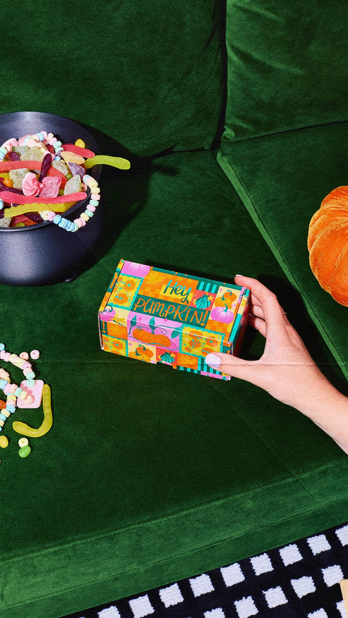 A hand reaches out to touch a brightly coloured "Hey Pumpkin!" gift box on a green velvet sofa, surrounded by a cauldron filled with sweets, candy necklaces, and an orange velvet pumpkin.