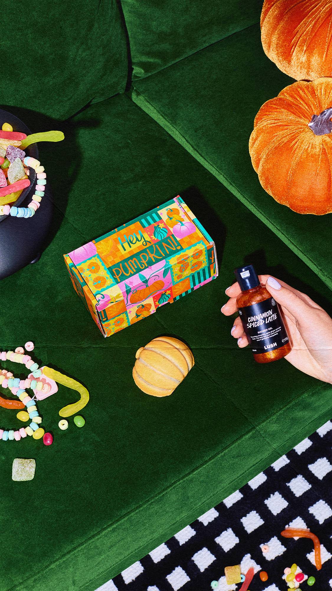 Top-down view on a green velvet sofa of a hand holding Cinnamon Spiced Latte shower gel beside a pumpkin bath bomb, a "Hey Pumpkin!" box, velvet pumpkins, and colourful sweets scattered around.