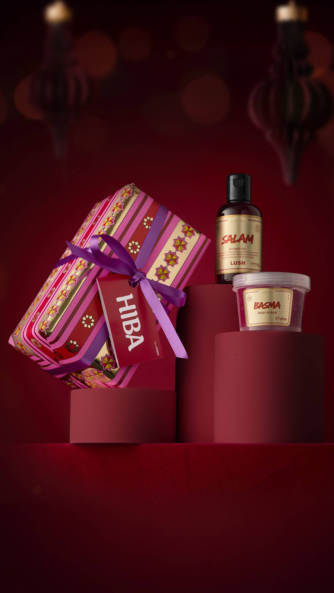 A selection of Lush Eid products, including a bottle of "Salam" shower gel, a pot of "Basma" body scrub, and the "Hiba" gift set, arranged on red pedestals. The gift is wrapped in gold and pink patterned paper with a purple ribbon.
