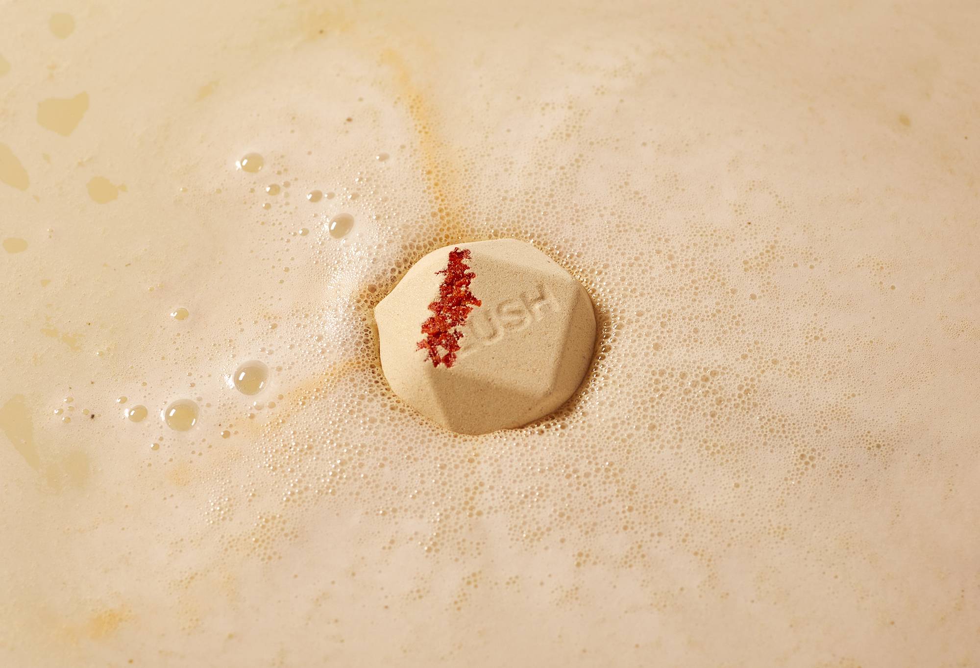 Hibana bath bomb sits on a sea of silky, creamy foam with red-coloured salt and "Lush" embossed on top.