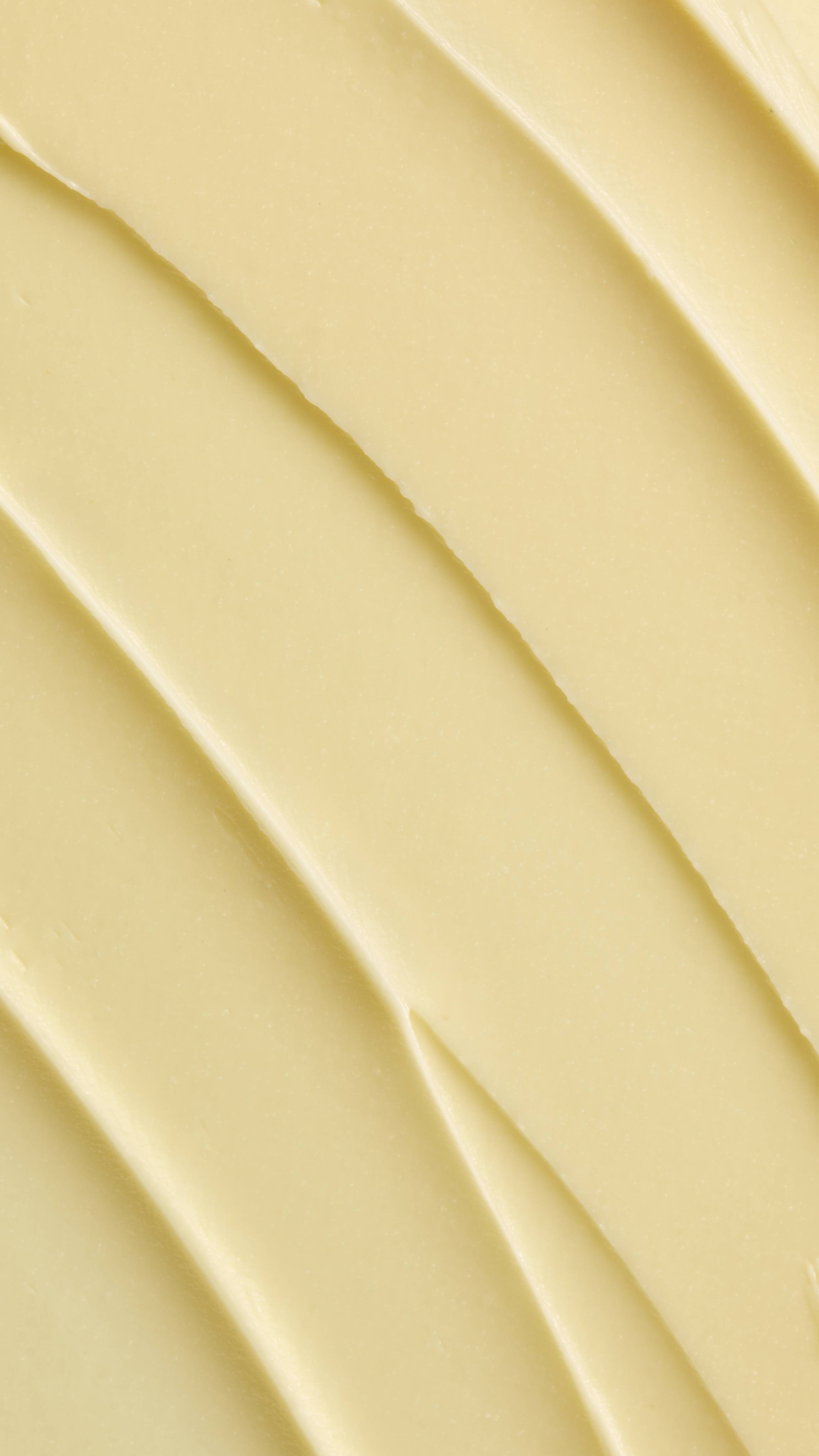 A super close-up image showing diagonal, overlapping smears of the thick, creamy, pale yellow Lush "Honey, Beeswax And Almond Oil" facial cleanser. The smooth product has a matte finish and subtle ridged textures.