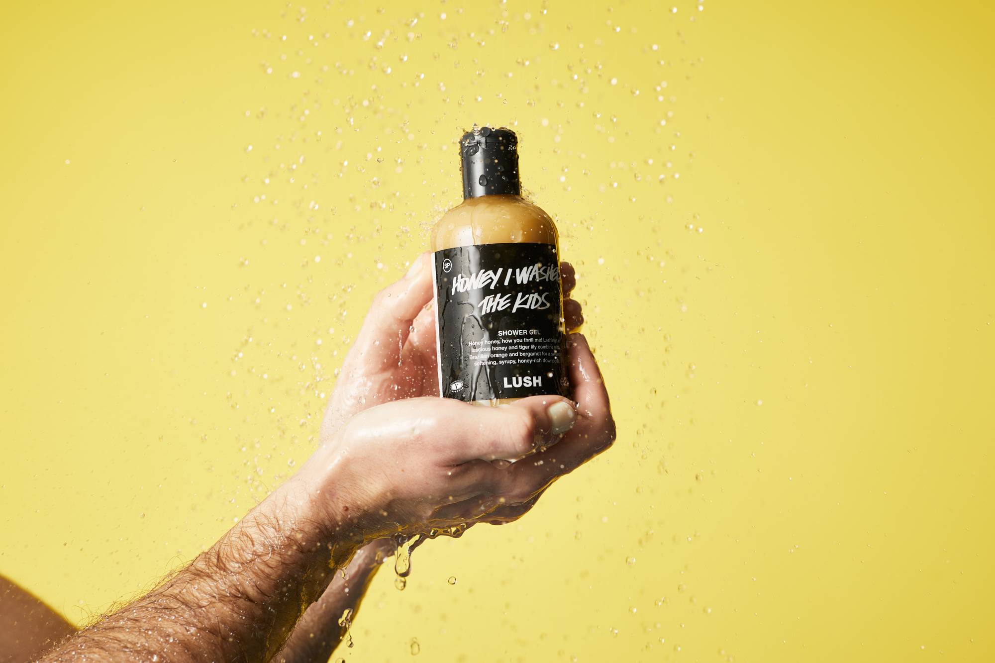 Shower water runs over a bright yellow background as we see a close-up of hands holding the shower gel bottle full of honey gel.