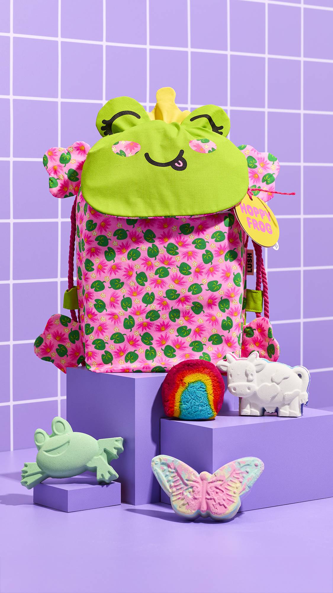 The Hoppy Frog gift backpack, patterned with pink lily pads, sits on purple pedestals. It is surrounded a Luna's butterfly bath bomb, a Toby Magic Cow bath bomb, a Mini Rainbow bubble bar, and a Hoppy Frog bath bomb against a purple tiled background.