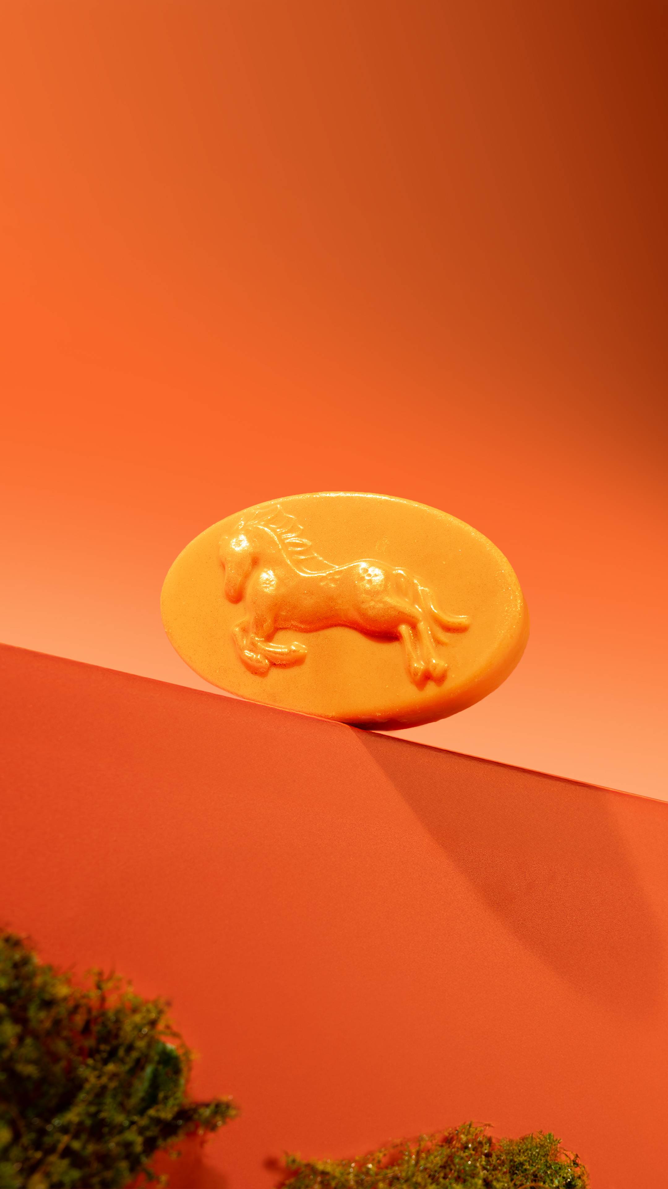 The oval HorsePower Massage Bar rests on a red geometric edge, showcasing a raised galloping horse design and shimmering Plastic Free Glitter above patches of green moss against an orange background.