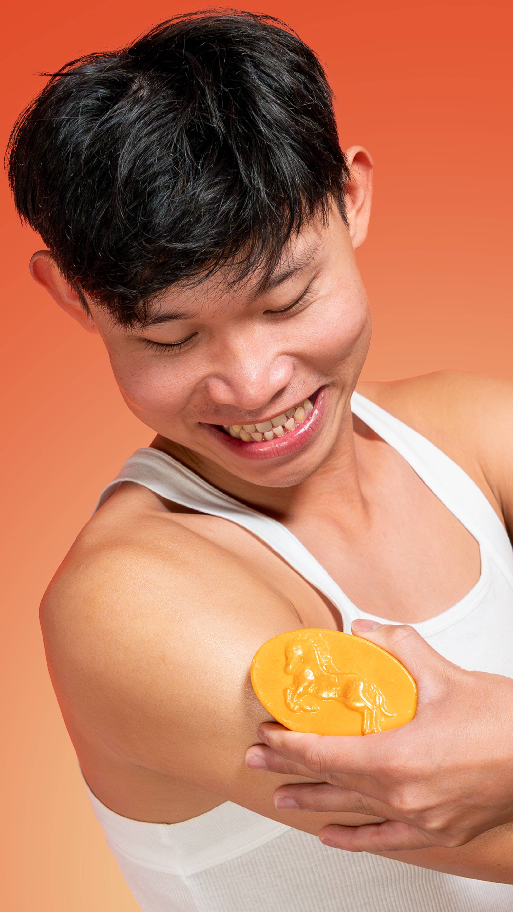 A smiling person with dark hair wears a white tank top and applies the golden HorsePower Massage Bar to their arm, showcasing the raised horse design against an orange background.