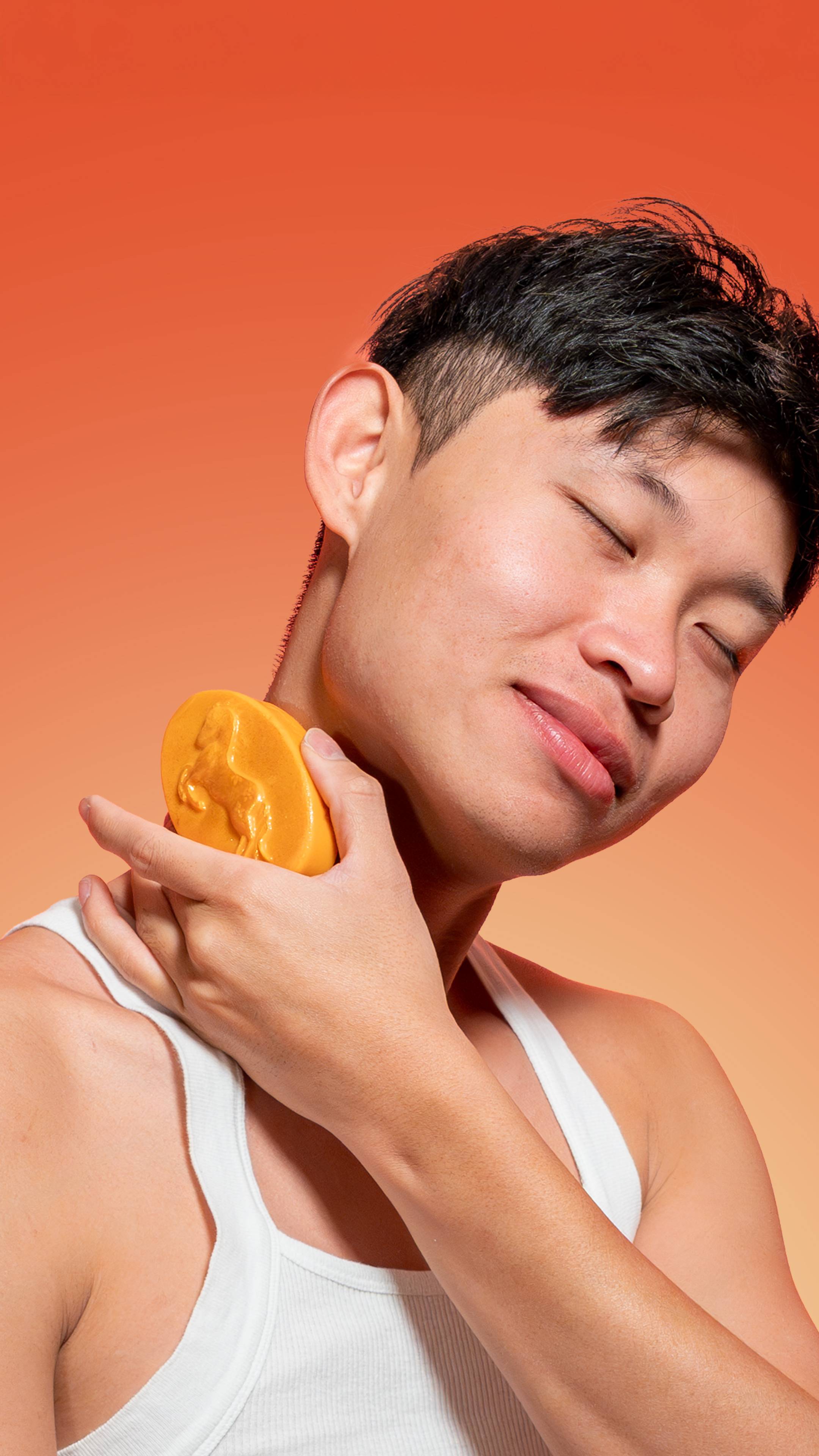 A smiling person with dark hair and closed eyes wears a white tank top and presses the golden HorsePower Massage Bar against their neck, the bar shimmering with Plastic Free Glitter against an orange background.