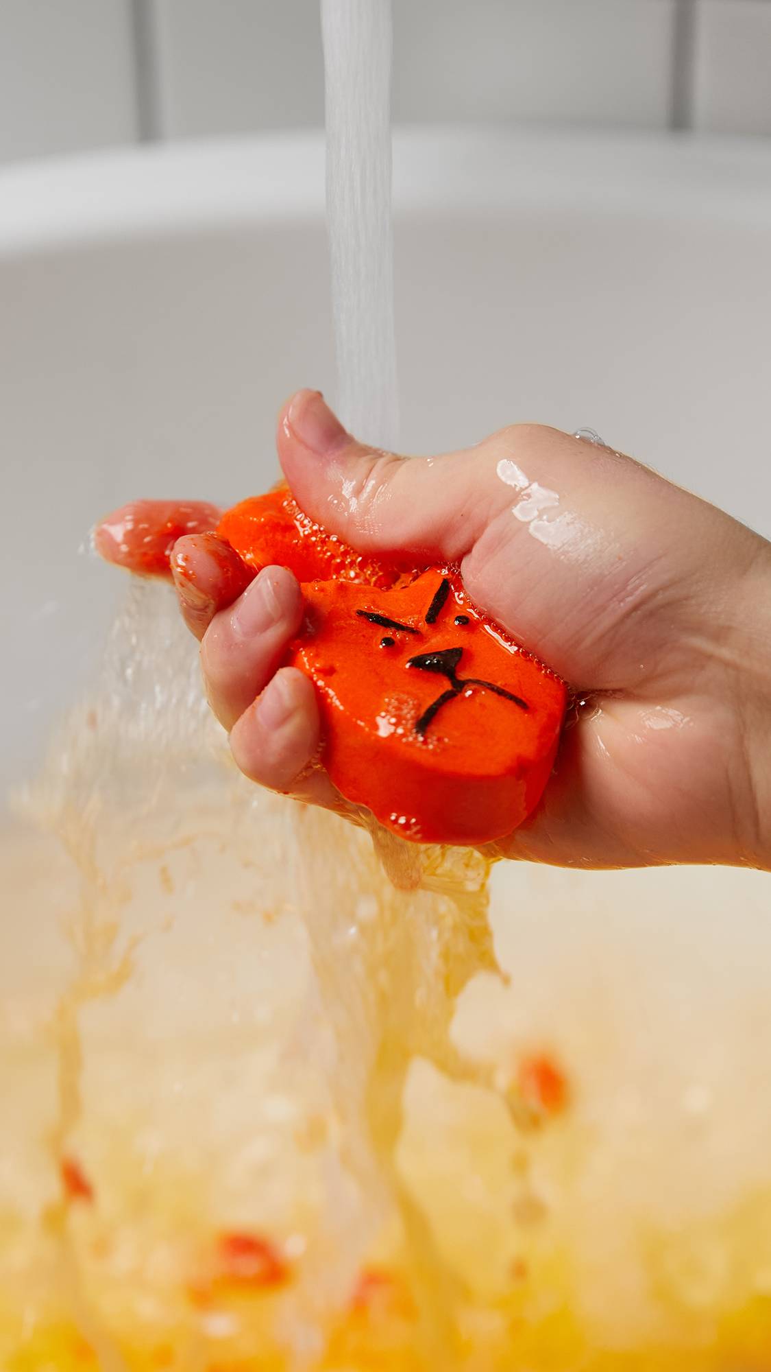 A close-up image of the model's hand crumbling the Hot Cross Bunny bubble bar under running water to create bubbly, orange waters below. 