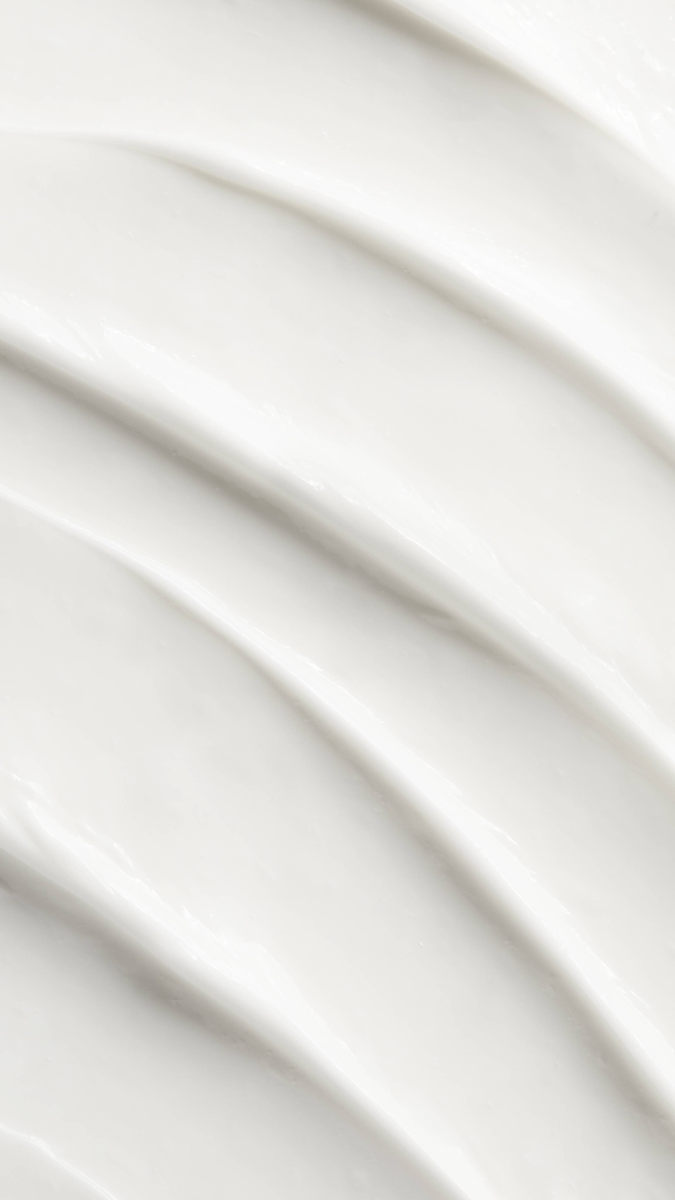 A close-up, aerial view of smooth, thick white  Hugs body lotion. The texture is rich and glossy, arranged in several diagonal, overlapping swoops that create soft ridges and valleys.