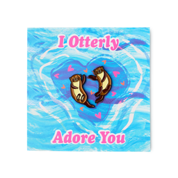 The "I Otterly Adore You" card features a blue swirling water design with pink text. Two brown otter enamel pins are attached to the center, surrounded by small pink hearts.