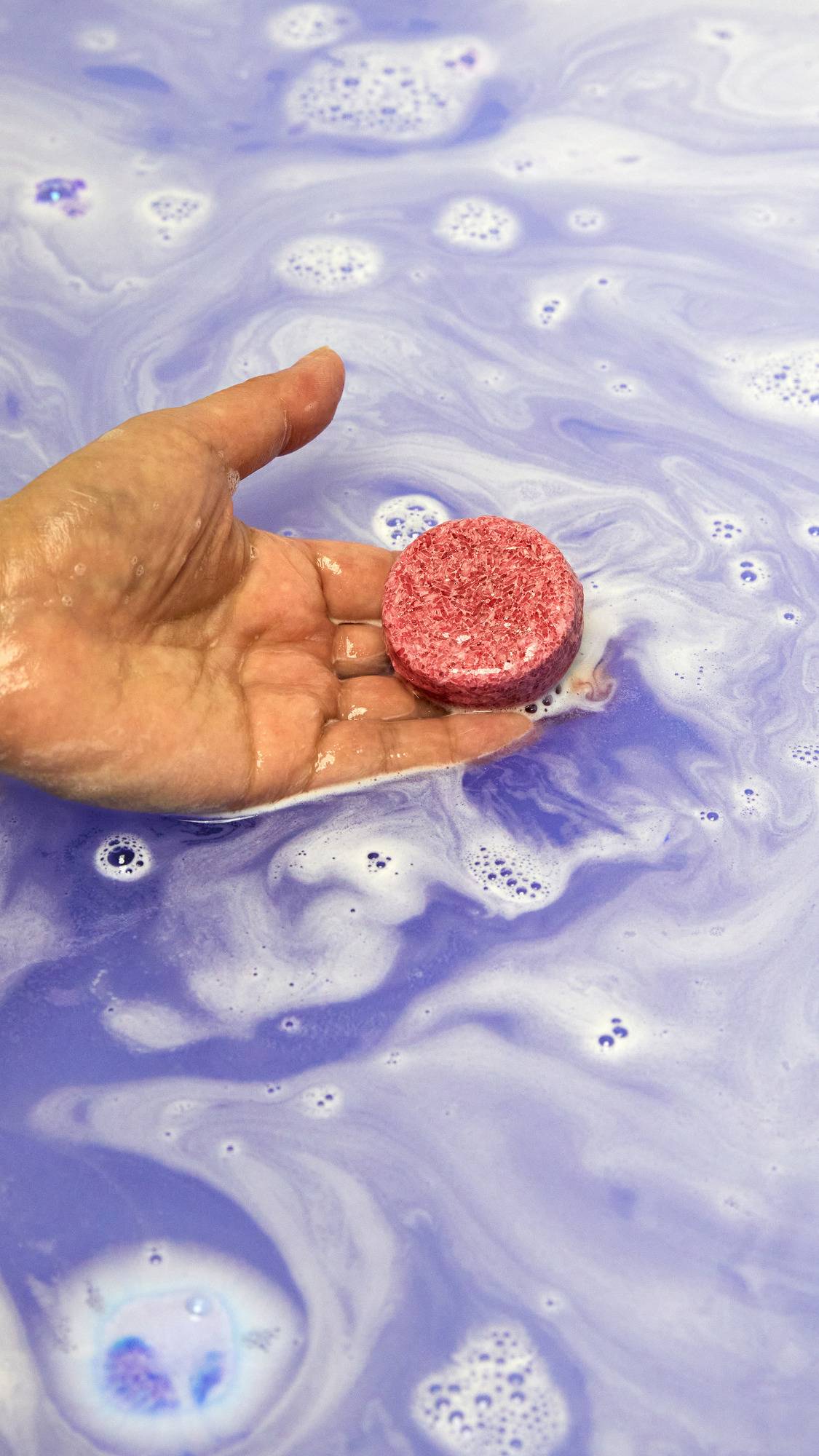 The Sleepy Bot bath bomb has completely dissolved in the water leaving behind a surprise mini reusable shampoo bar held by the model above pastel purple waters. 