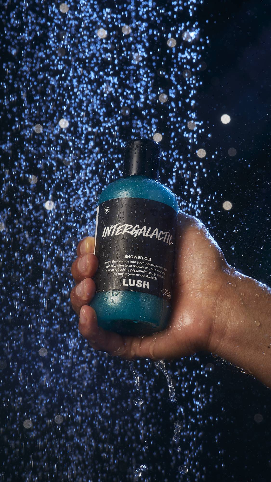 Image shows a close-up of the model's hand holding the Intergalactic shower gel bottle under blue-tinted shower water. 