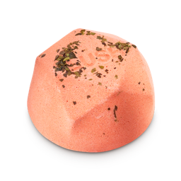 Hinamatsuri. A peach, half-sized bath bomb in a geometric dome shape. The flat top is sprinkled with dried green botanical leaves and features embossed "LUSH" text.