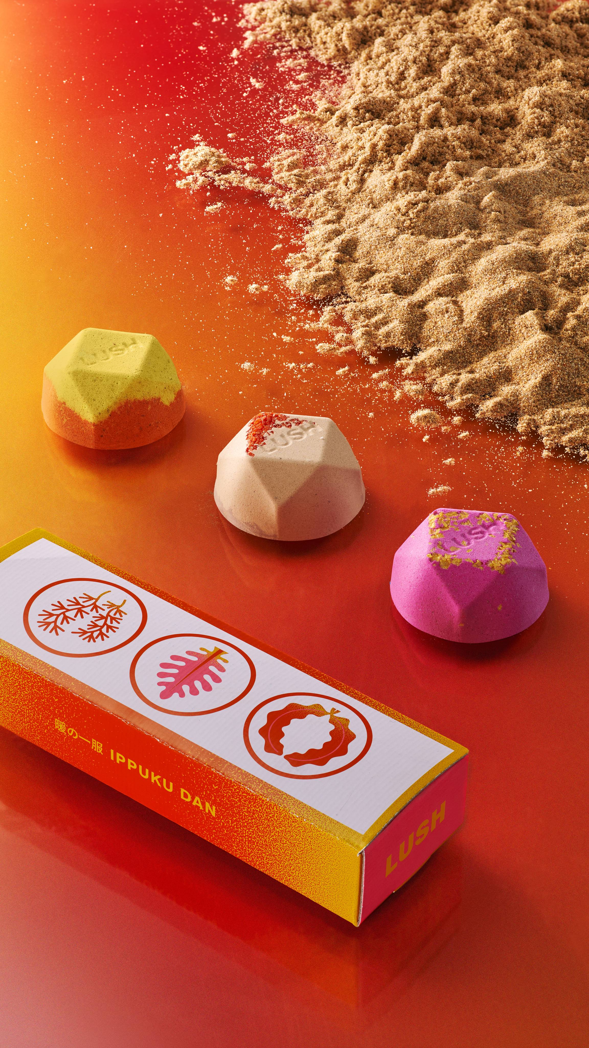 Three ippuku warming bath bombs lay in a line with sand on one side, the box on another on top of an orange-yellow gradient.