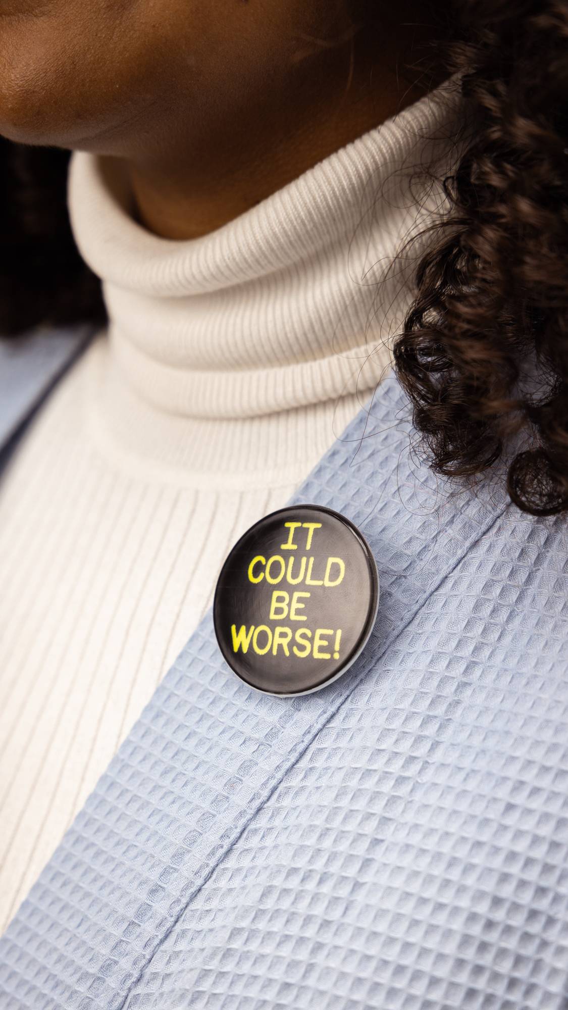 Model is wearing a white turtle neck and a pale blue jacket. They are wearing the "It Could Be Worse" badge on their lapel.