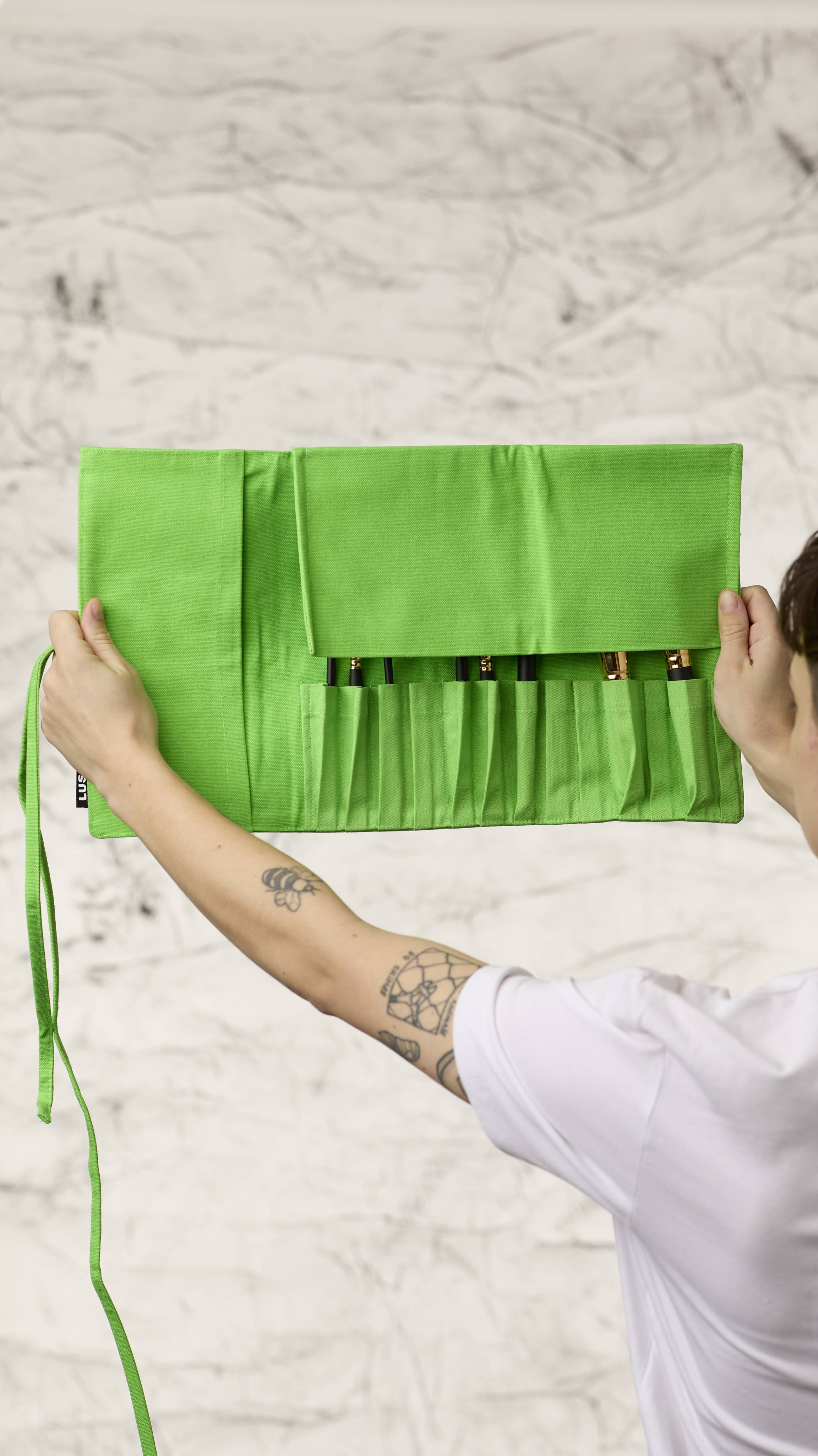 The image shows a blurred, marble background. Two hands are holding a fluorescent green roll-up makeup brush wrap as it shows eight different kinds of makeup brushes in the slots at the bottom with the top flap covering the brush heads.