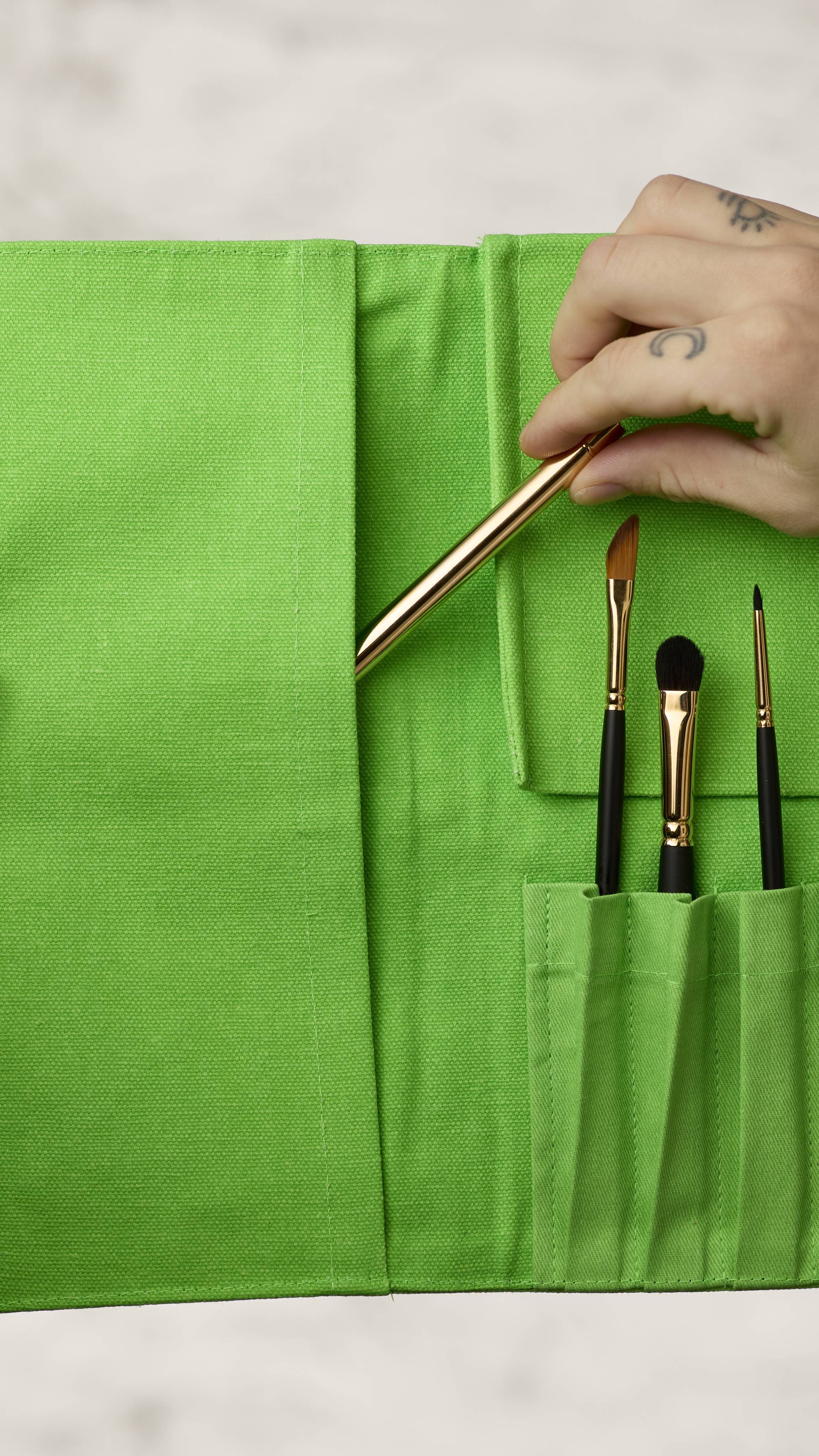 The image shows a blurred, marble background. A hand is reaching into the left side panel of the fluorescent green roll-up makeup brush wrap to access extra brush storage and is pulling out a long, thin makeup brush.