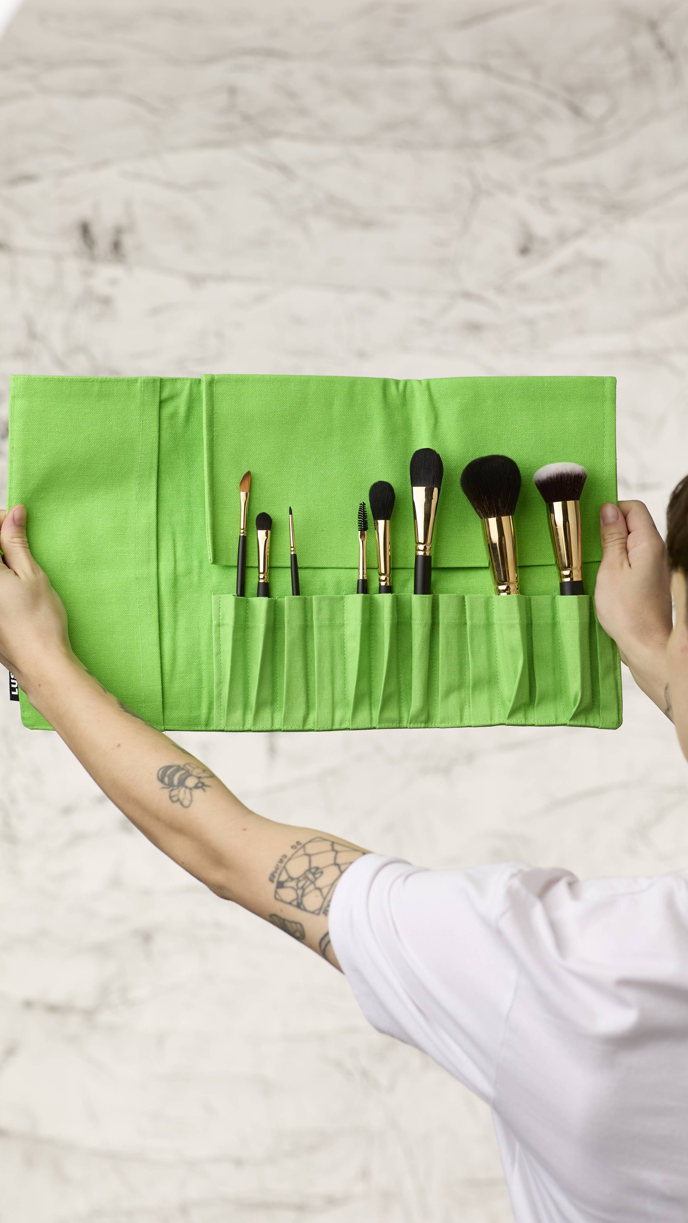The image shows a blurred, marble background. Two hands are holding a fluorescent green roll-up makeup brush wrap as it shows eight different kinds of makeup brushes in the slots at the bottom.