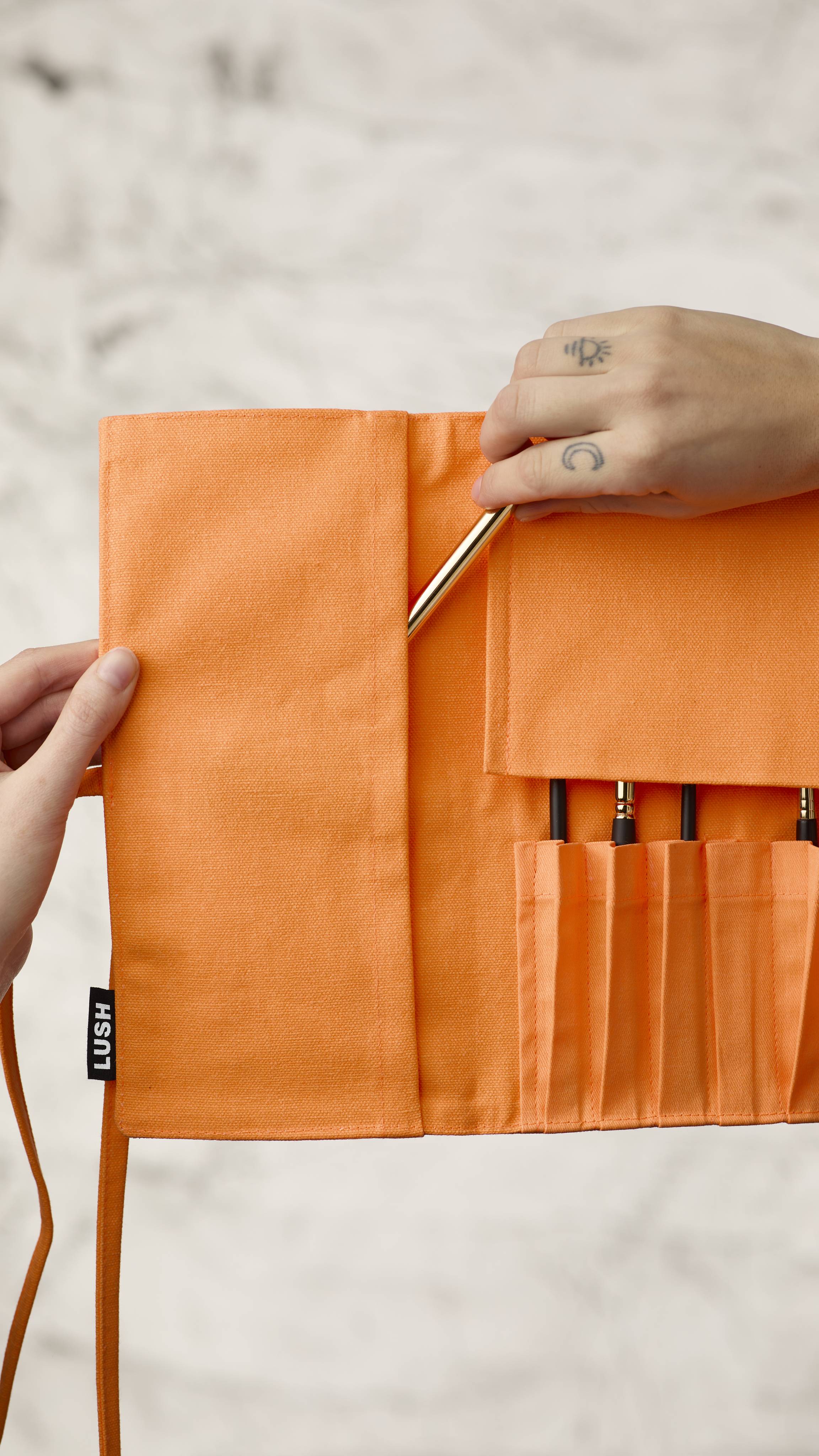The image shows a blurred, marble background. A hand is reaching into the left side panel of the fluorescent orange roll-up makeup brush wrap to access extra brush storage and is pulling out a long, thin makeup brush.