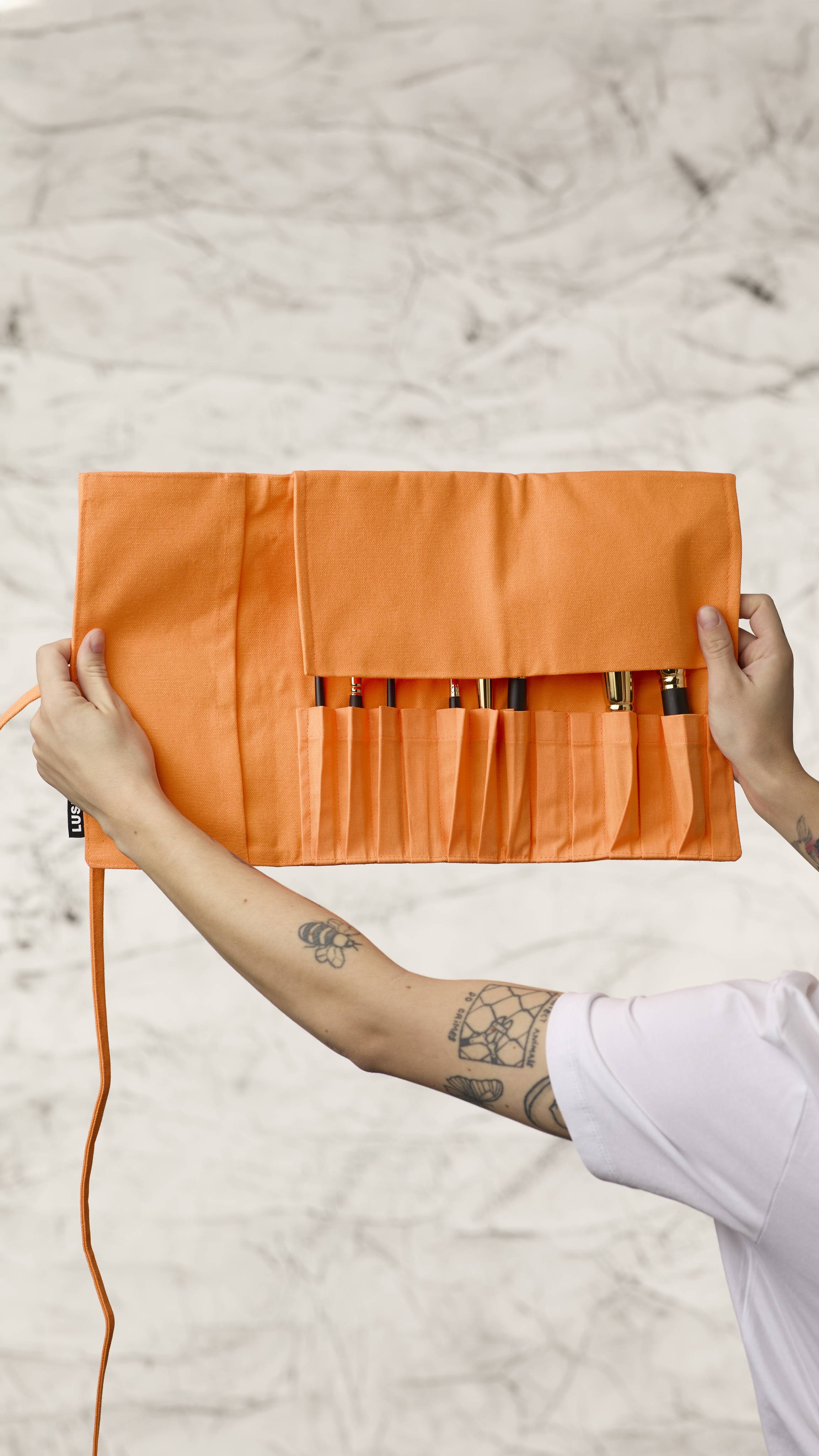 The image shows a blurred, marble background. Two hands are holding a fluorescent orange roll-up makeup brush wrap as it shows eight different kinds of makeup brushes in the slots at the bottom with the top flap covering the brush heads.