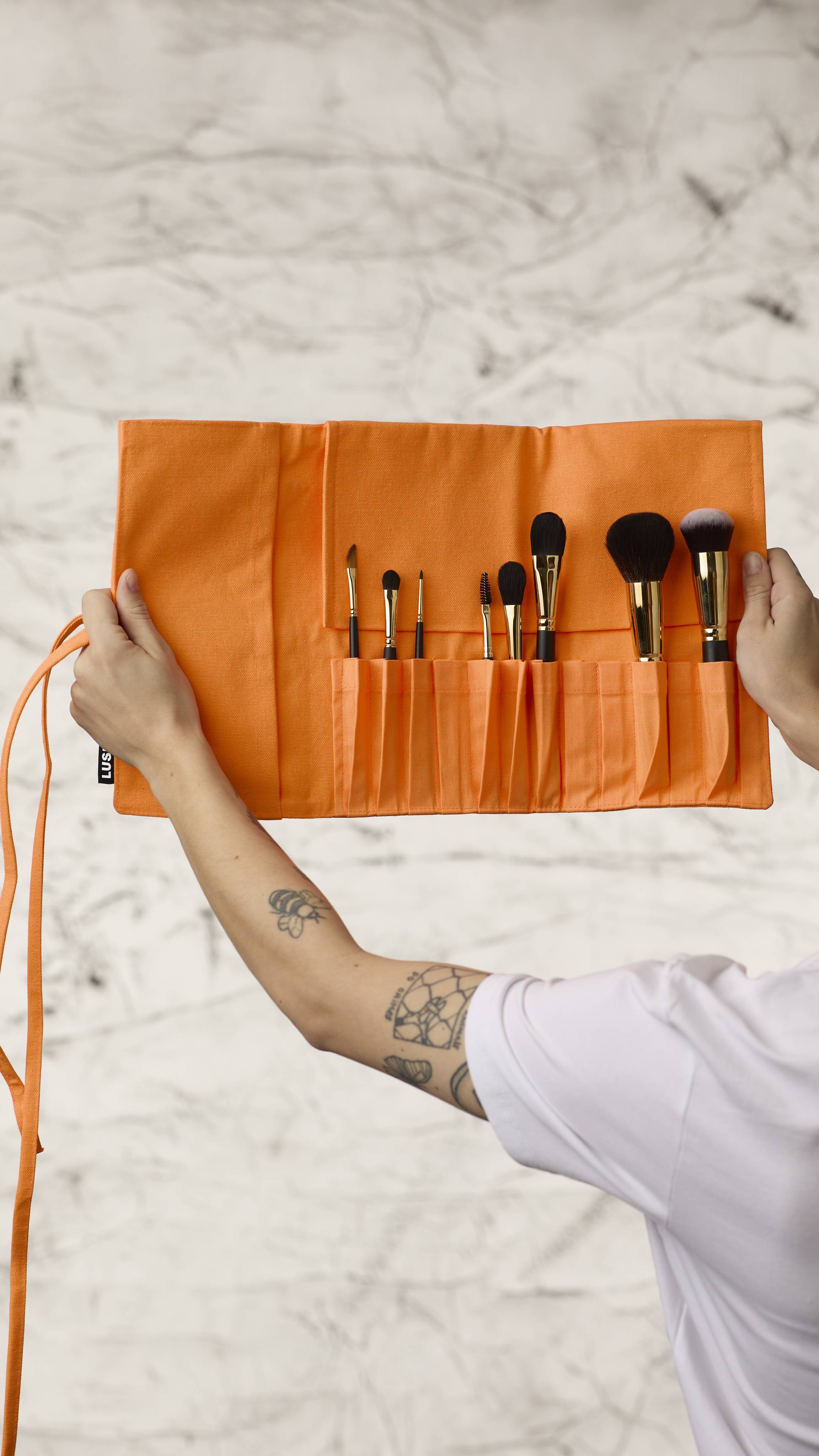 The image shows a blurred, marble background. Two hands are holding a fluorescent orange roll-up makeup brush wrap as it shows eight different kinds of makeup brushes in the slots at the bottom.