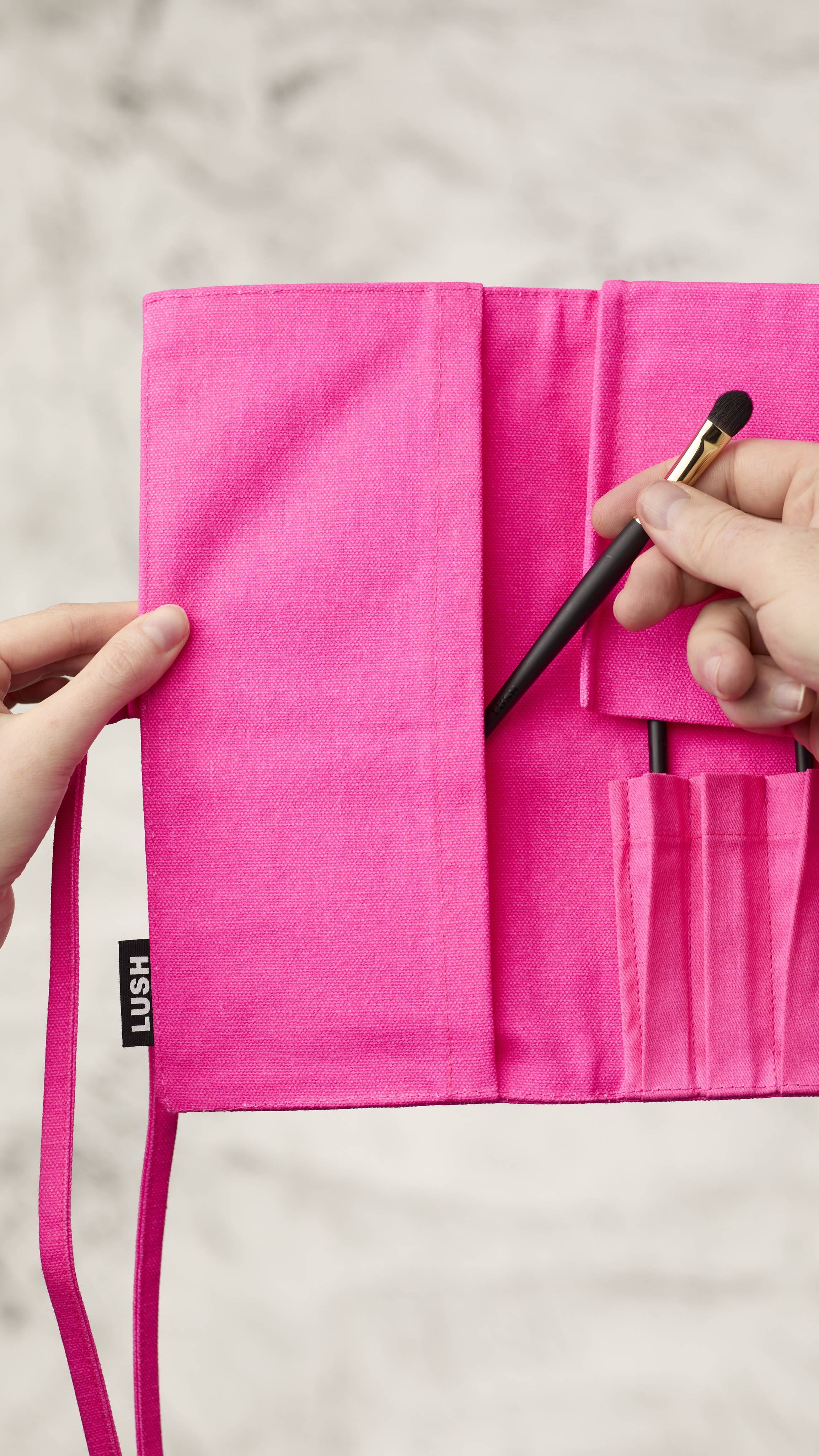The image shows a blurred, marble background. A hand is reaching into the left side panel of the fluorescent pink roll-up makeup brush wrap to access extra brush storage and is pulling out a long, thin makeup brush.