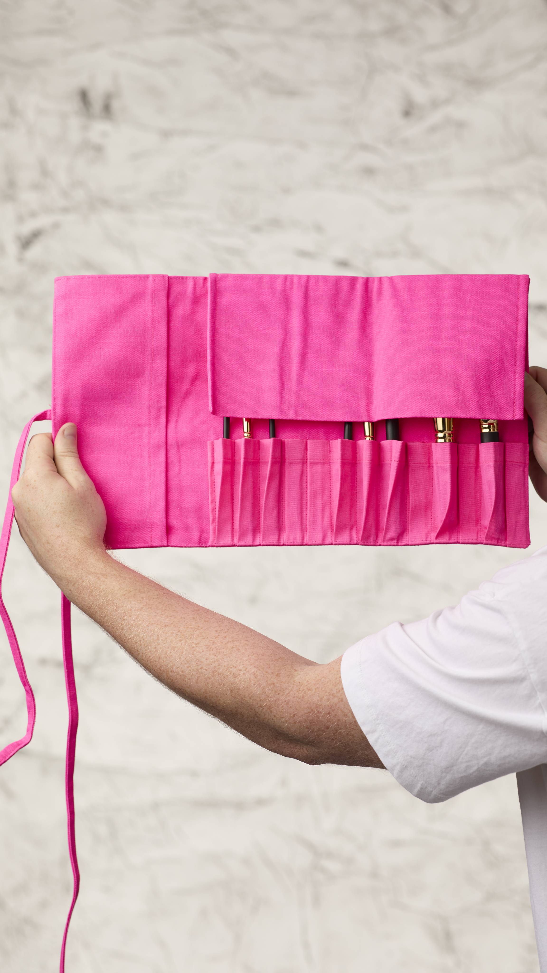 The image shows a blurred, marble background. Two hands are holding a fluorescent pink roll-up makeup brush wrap as it shows eight different kinds of makeup brushes in the slots at the bottom with the top flap covering the brush heads.