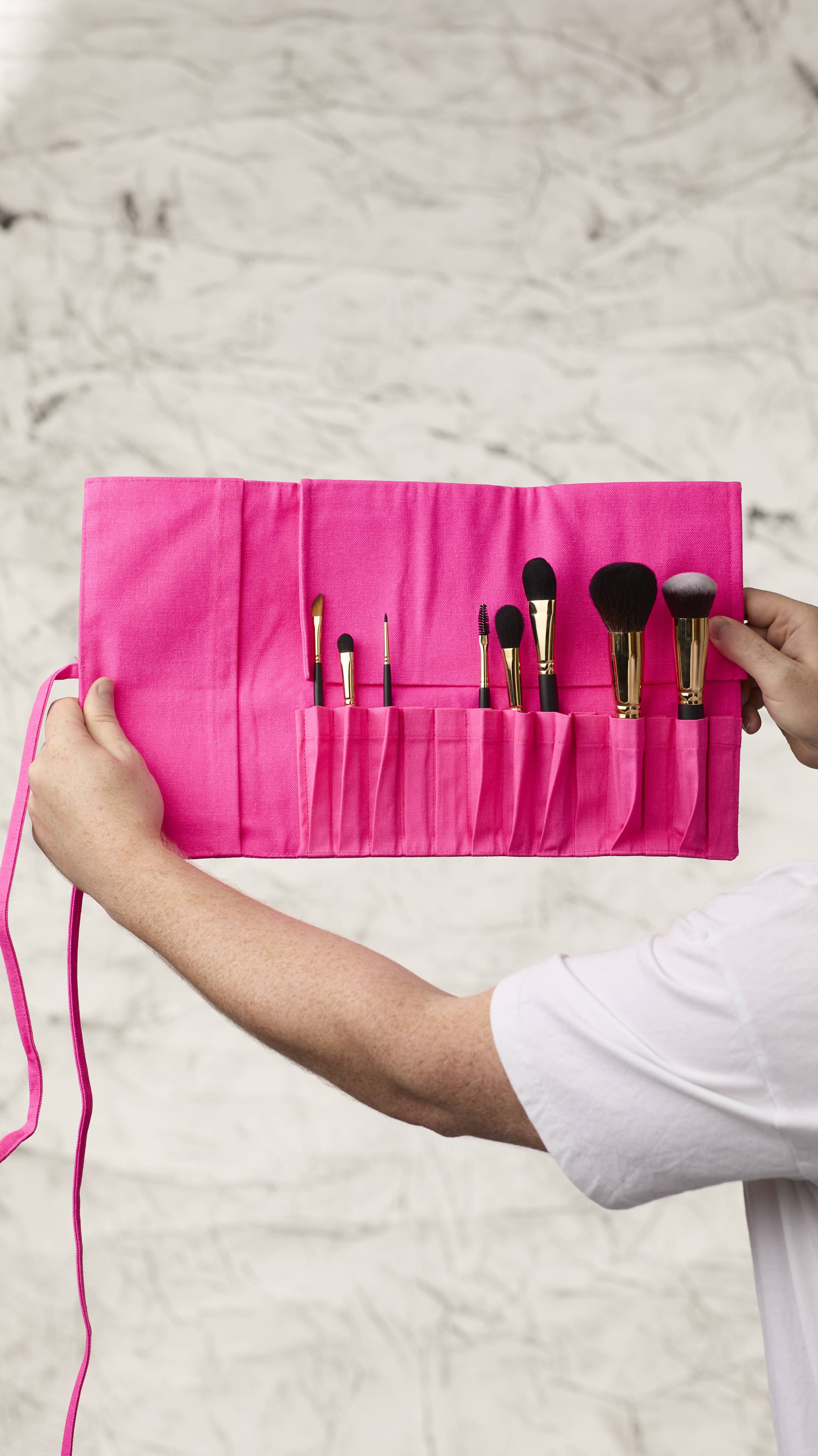 The image shows a blurred, marble background. Two hands are holding a fluorescent pink roll-up makeup brush wrap as it shows eight different kinds of makeup brushes in the slots at the bottom.