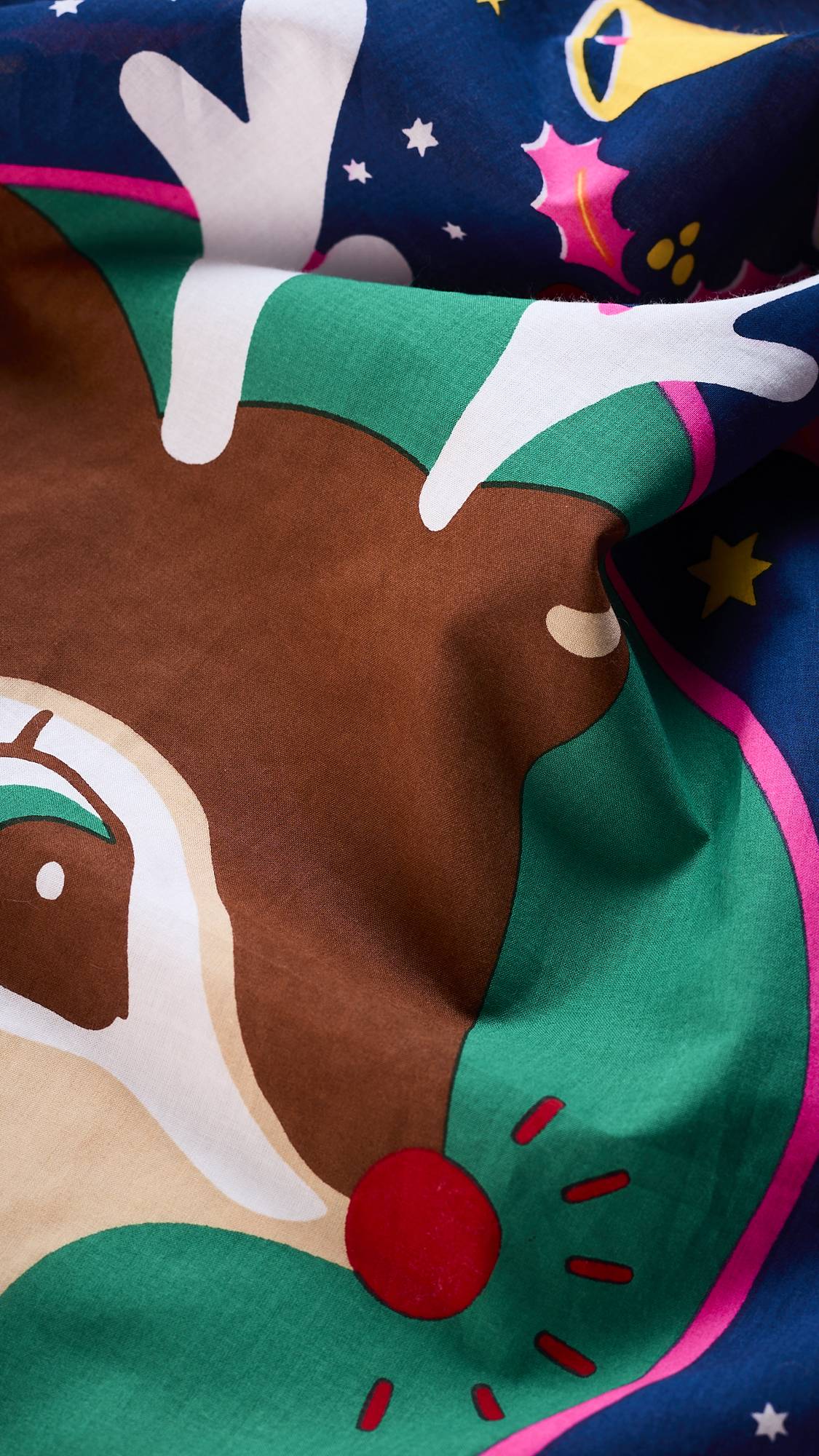 A close-up of the "Christmas Deer" Knot Wrap, a reusable fabric from Lush. The pattern features a cartoon reindeer with a bright red nose and white antlers surrounded by gold stars, green holly leaves, and pink bells.