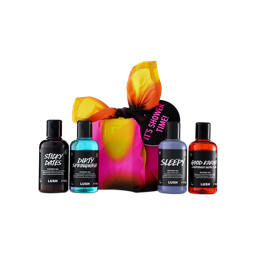 A colourful tie-dye knot-wrapped gift with an "IT'S SHOWER TIME!" tag sits against a black background, flanked by four Lush shower gels: Sticky Dates, Dirty Springwash, Sleepy, and Good Karma.