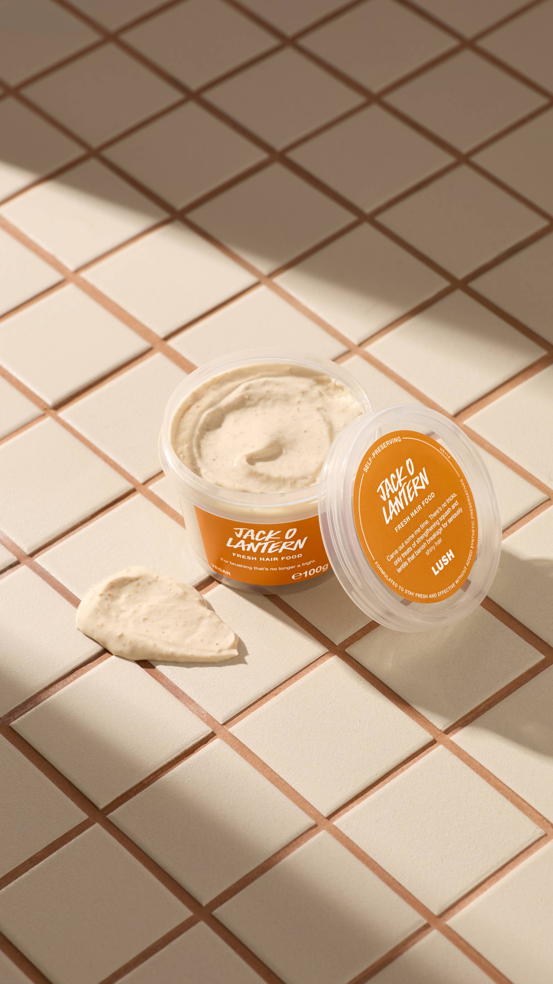 An open clear pot of Lush "Jack O Lantern Fresh Hair Food" filled with beige, textured cream sits on neutral tiled surfaces beside its clear lid with an orange label, accompanied by a small dollop of the product.