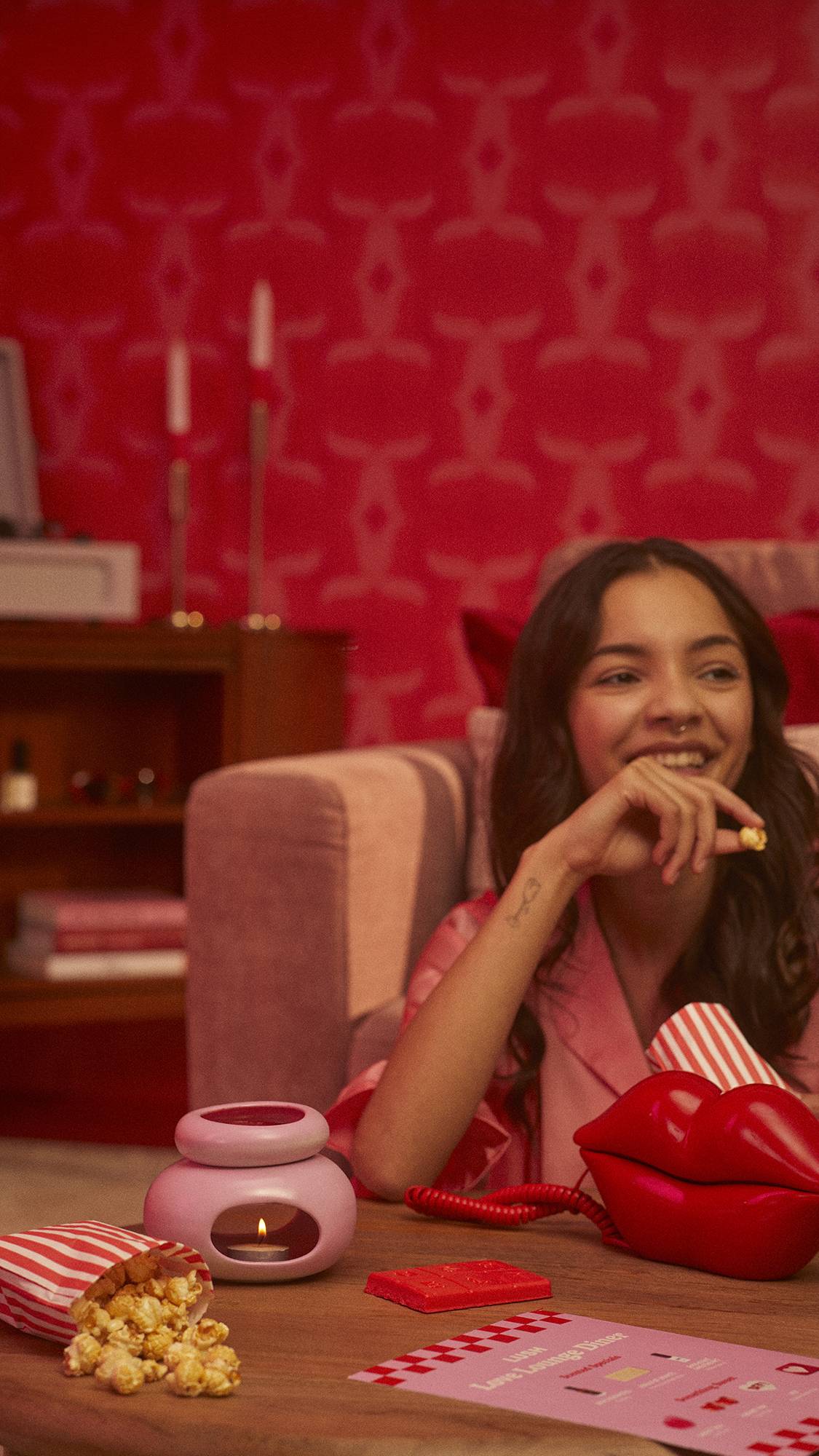The model. is in a red-themed room, eating popcorn as the product slowly melts in the wax burner.
