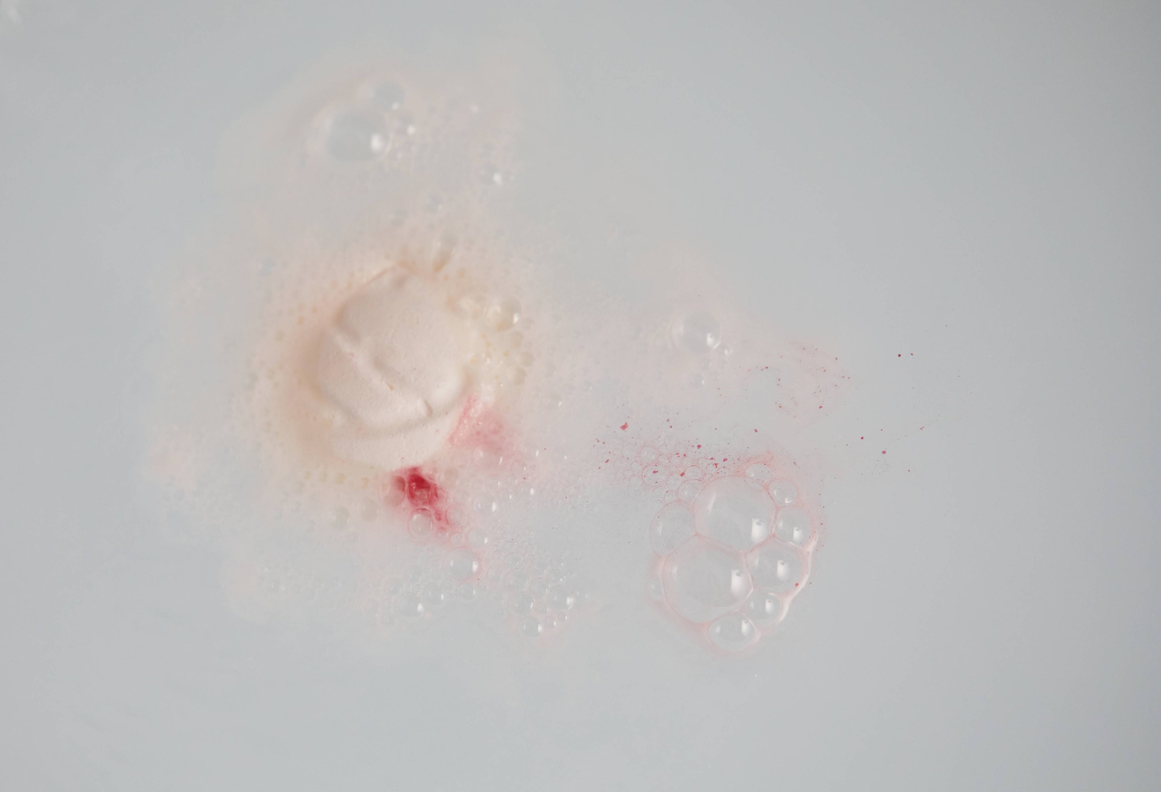 Bath bomb slowly dissolves in the bathwater creating subtle, creamy swirls of velvety water as the red starts to seep out.