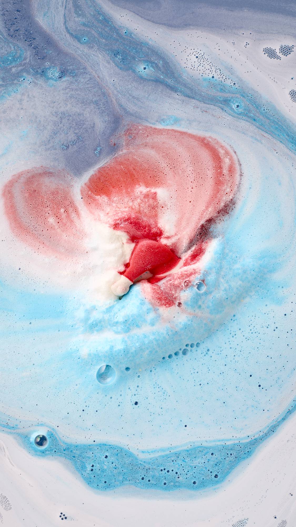 A Santa-shaped bath bomb dissolves in bathwater, releasing a fan-like spread of vivid red and white foam surrounded by bright sky-blue fizz and swirling icy blue water.