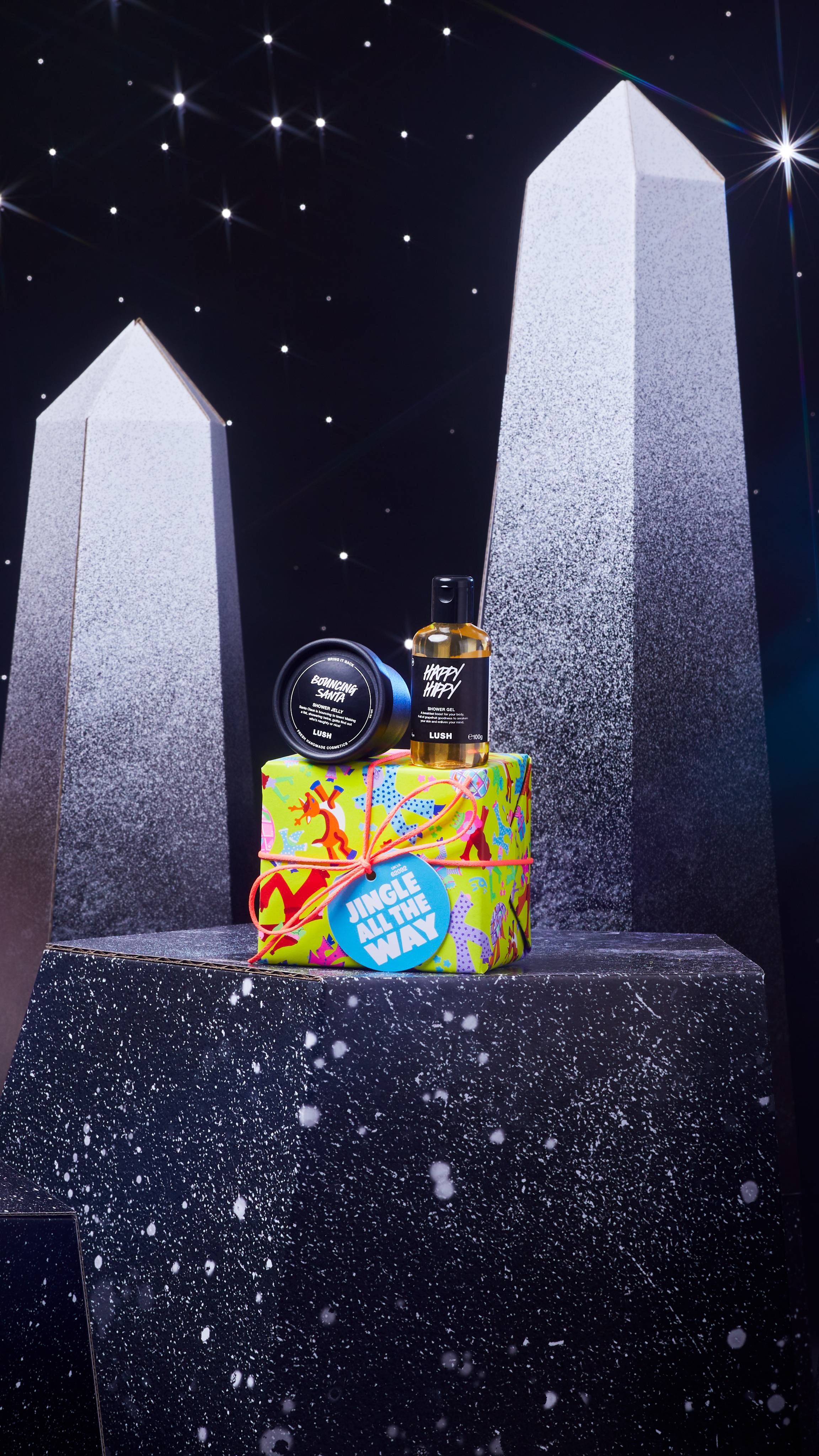 Jingle All The Way gift box and two shower products are on a dark chromatic platform among geometric shapes. 