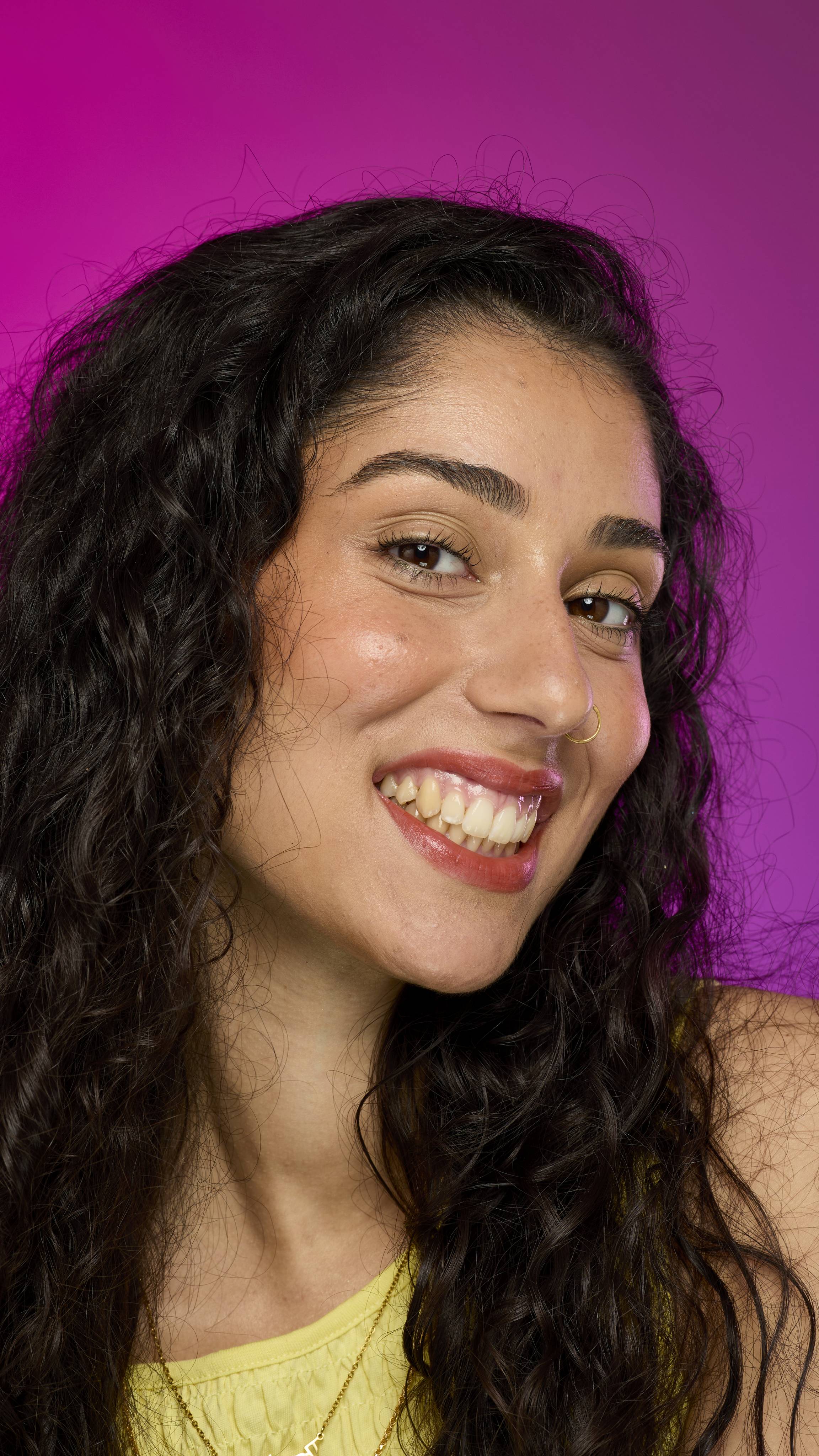 A smiling person with long, dark hair, wearing a yellow top, is shown from the chest up. They are on a vibrant pink background, showing off the rusted bronze-coloured Jumping Jack lipstick with a subtle glittering shimmer. 
