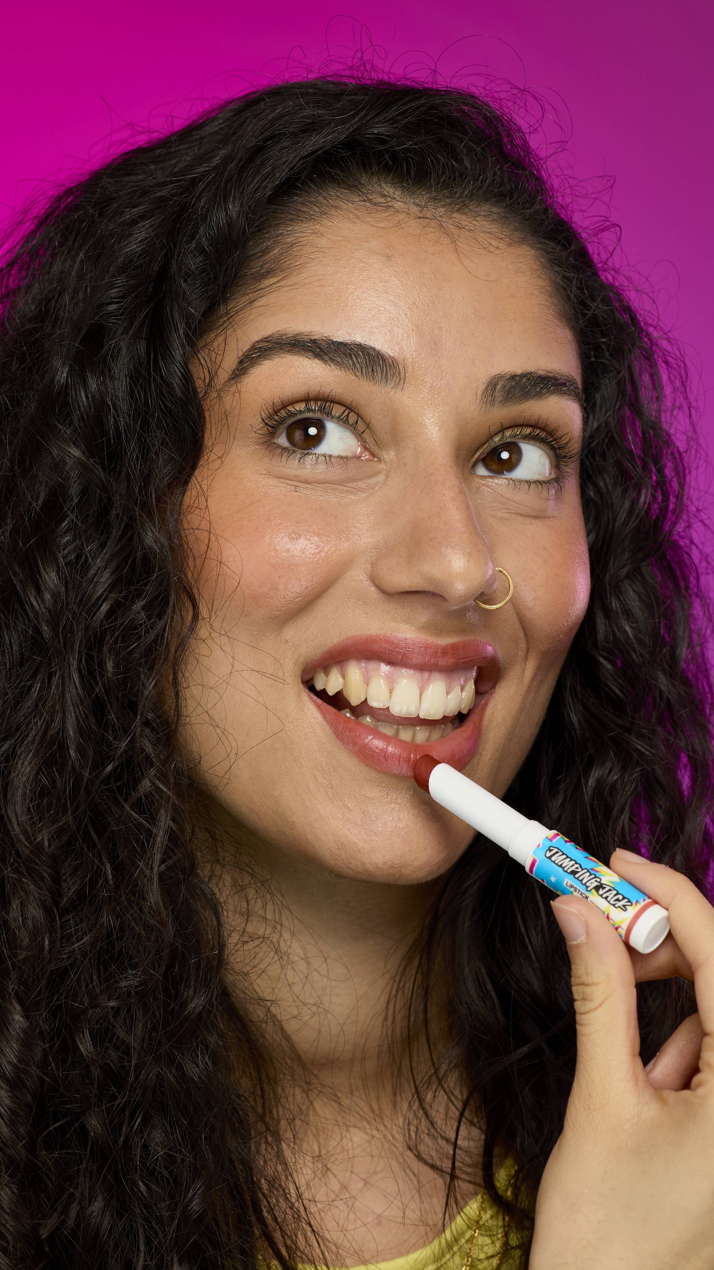A model with long, dark hair, wearing a yellow top, is shown from the chest up. They are on a vibrant pink background as they apply the twist-up Jumping Jack lipstick. 