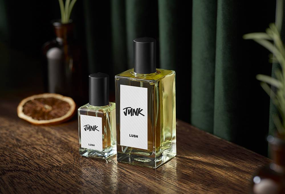 Perfume stands on a dark wood surface. A dried orange and greenery in a vase can be seen  against a dark green velvet curtain.  
