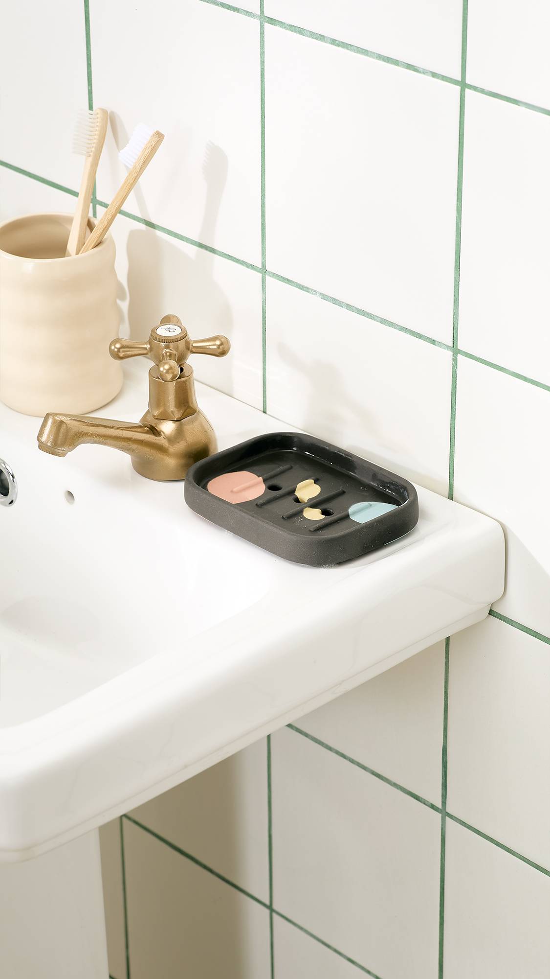 A dark grey ceramic soap dish featuring pastel spots sits beside a brass tap on a white pedestal sink, next to a ceramic holder with wooden toothbrushes set against a white tiled wall.   