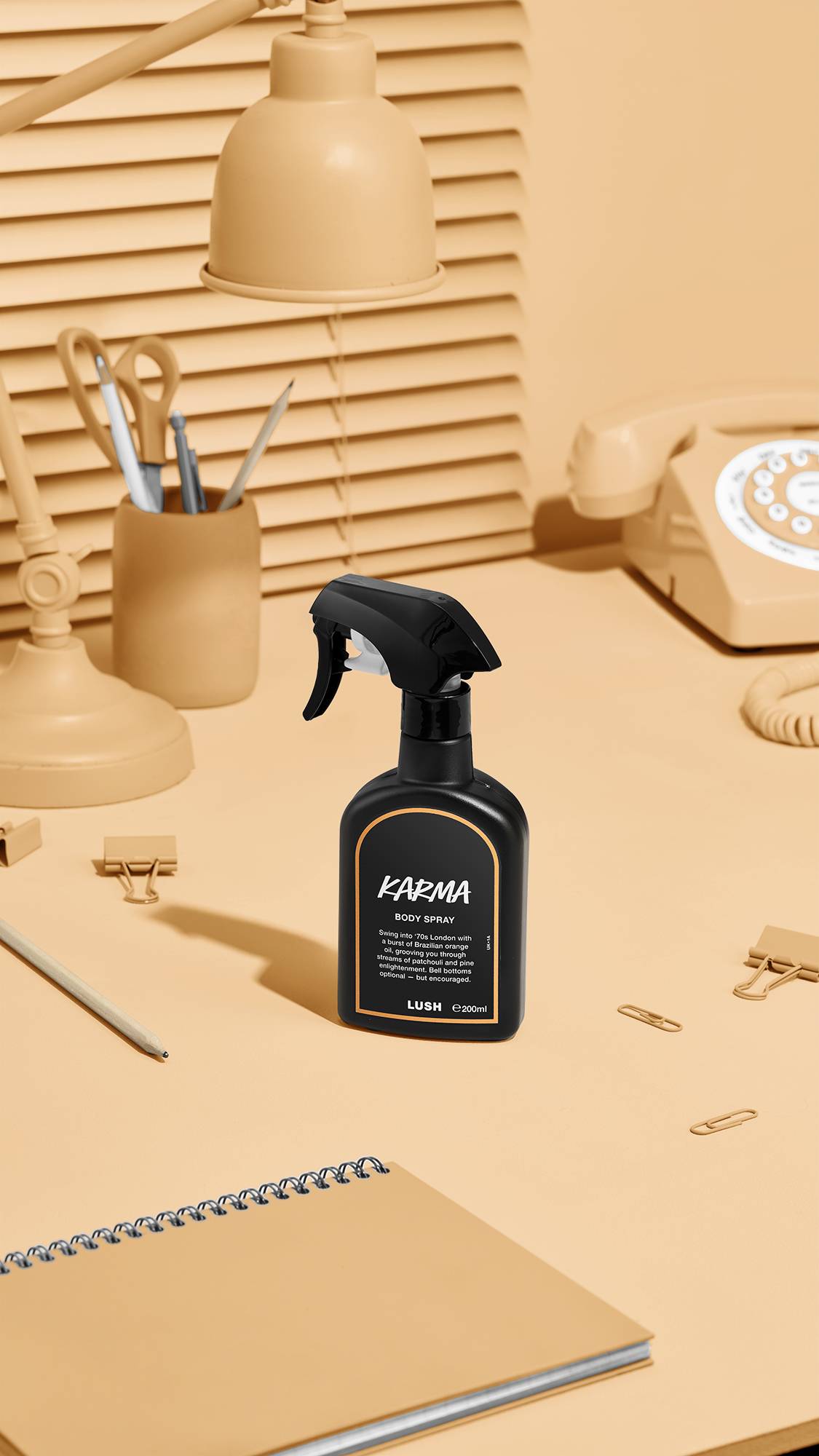 The image shows a black LUSH 'Karma' body spray bottle with an orange rimmed label on a beige desk. Nearby sit a matching desk lamp, a rotary phone, stationery, and a notebook, against window blinds.