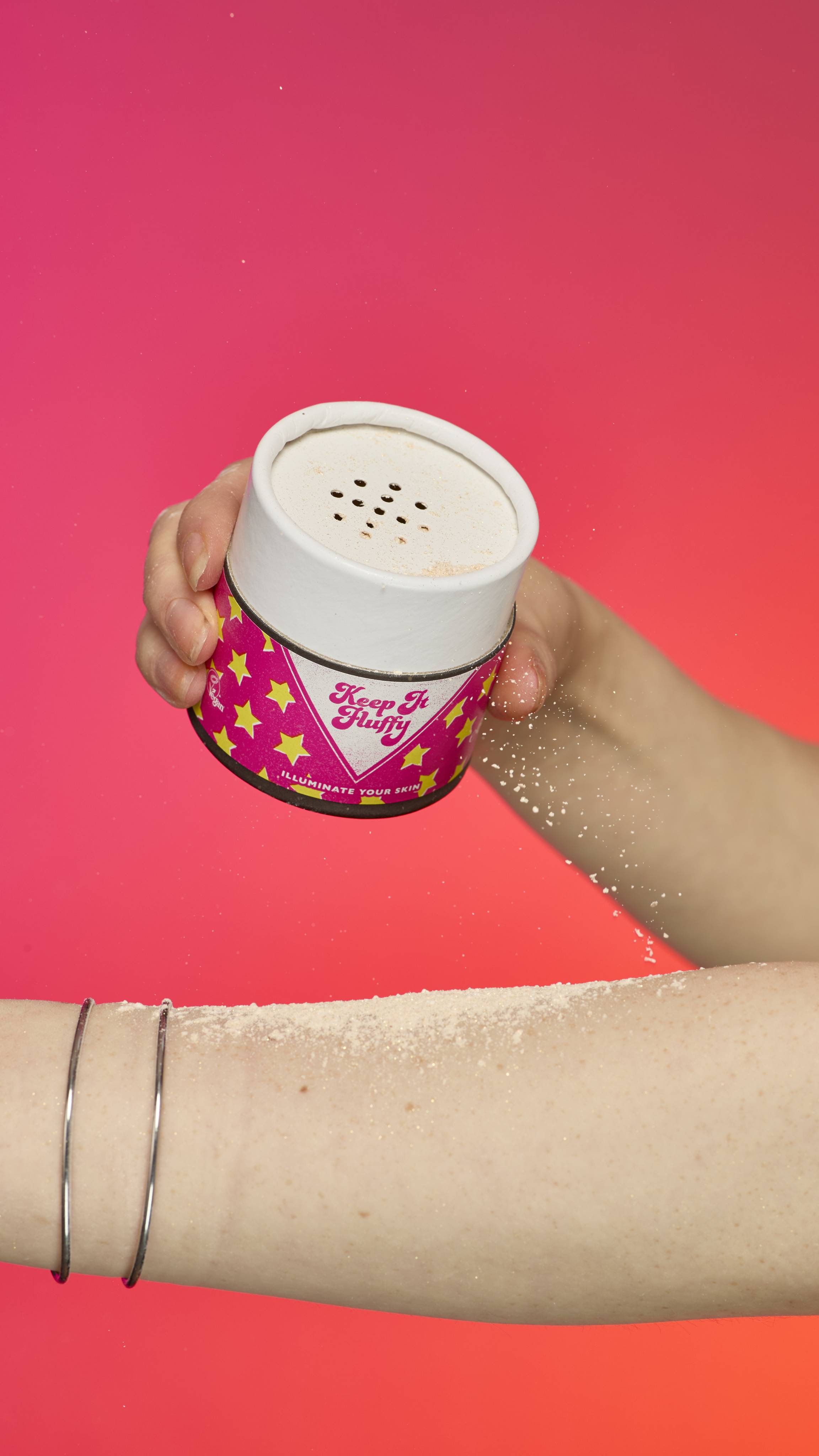 The image shows a hand holding a cylindrical shaker container of Keep It Fluffy sparkly body powder. They sprinkle the powder onto their forearm. The wrist has two simple silver bangle bracelets, and the background is a gradient of pink and orange.