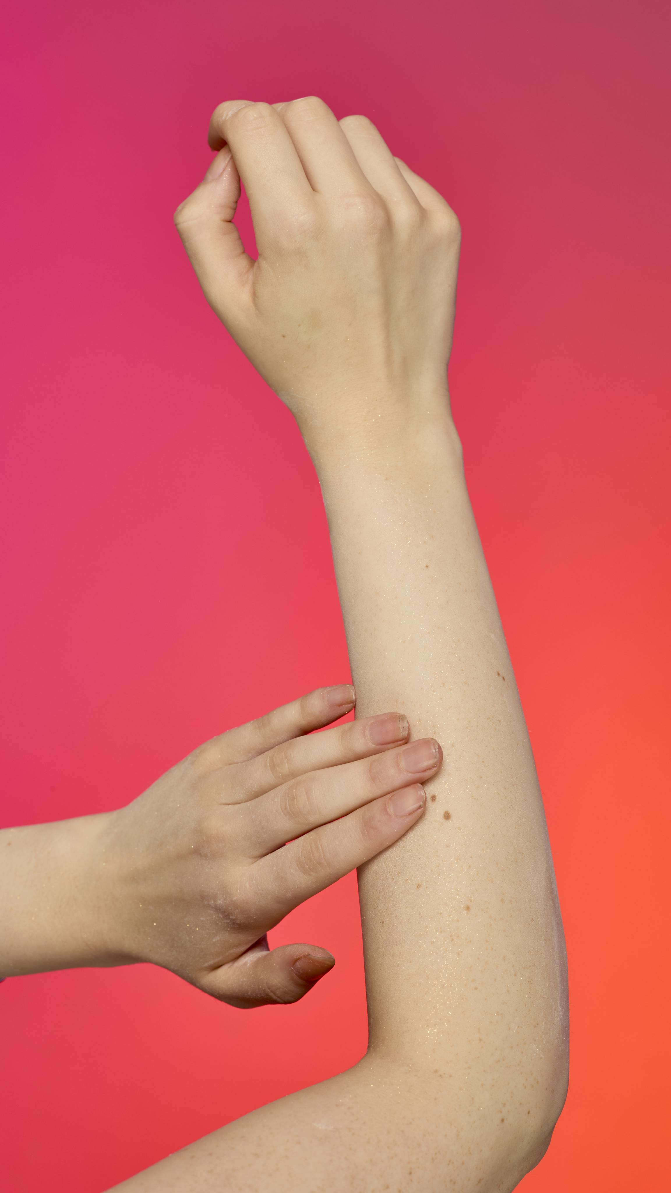 The image shows a light-toned hand reaching in to gently touch a forearm that is dusted with Keep It Fluffy sparkly body powder. The powder creates a subtle glitter effect on a pink and orange gradient background.