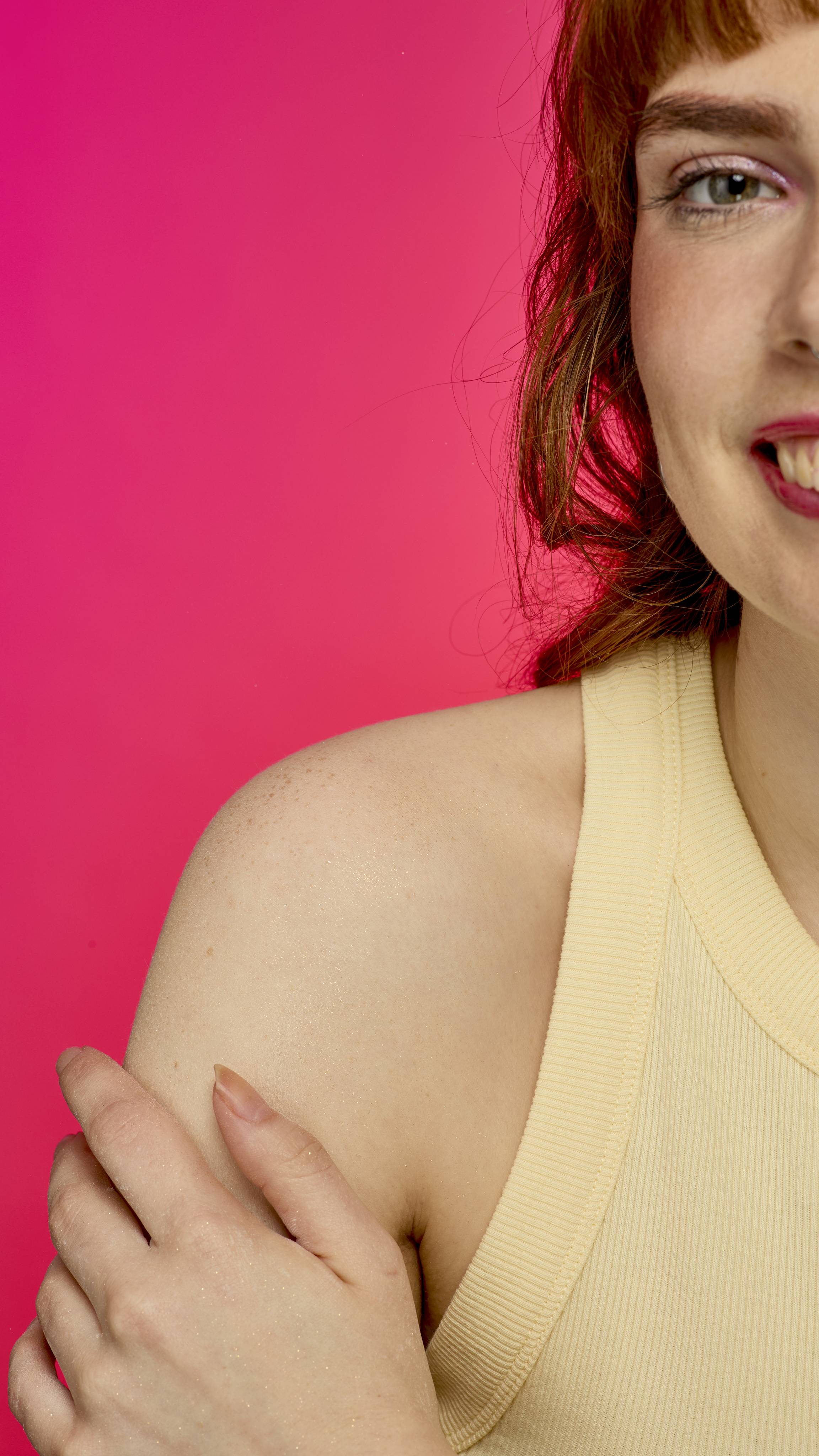 A smiling person with reddish hair and a yellow tank top is shown from the shoulder up. Their bare shoulder and upper arm are dusted with the Keep It Fluffy sparkly body powder. They are holding their arm, and the background is a solid bright pink.