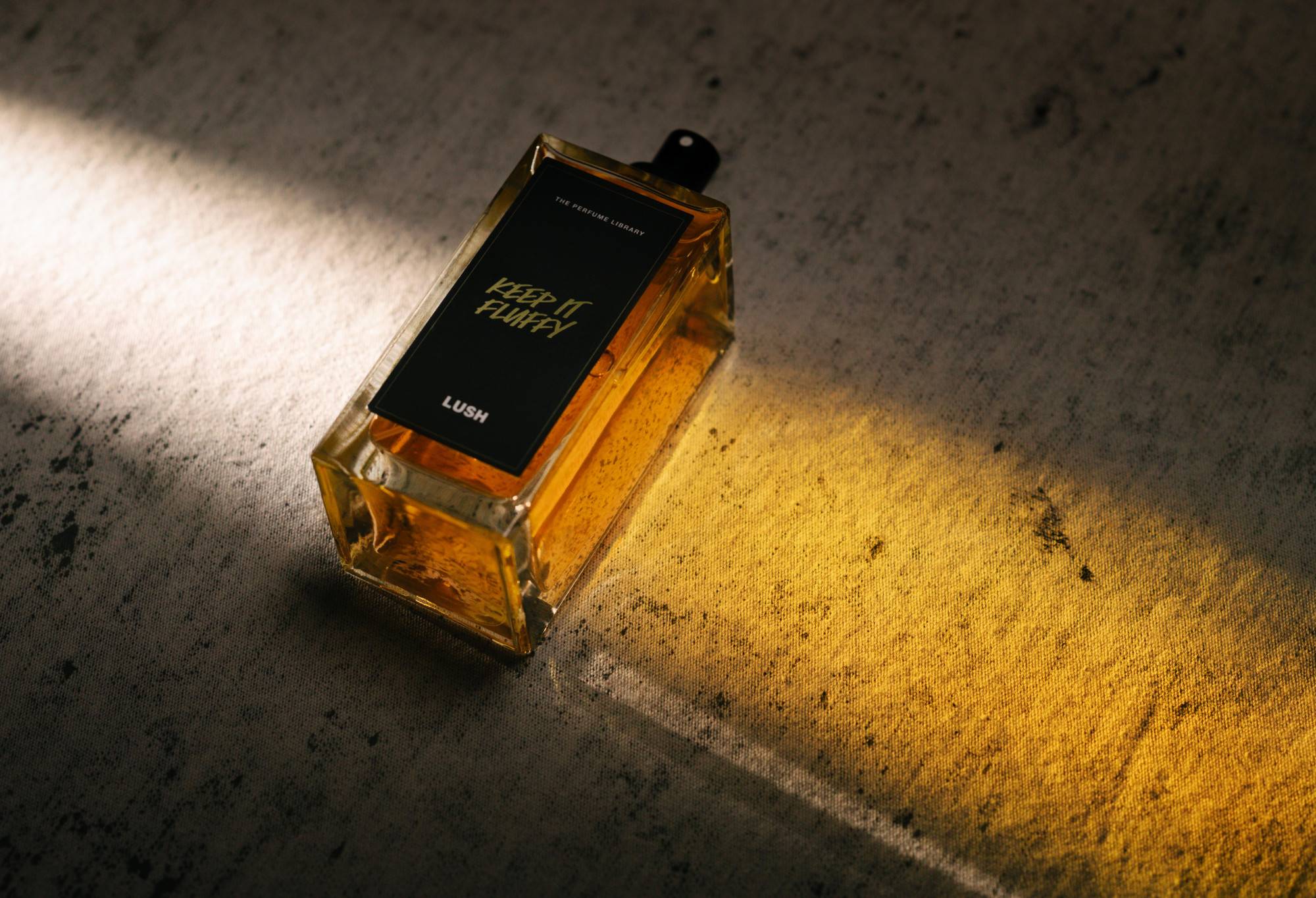 Keep It Fluffy, a bottled, black label perfume, lies flat, with light shining through it, creating an amber cast of light.