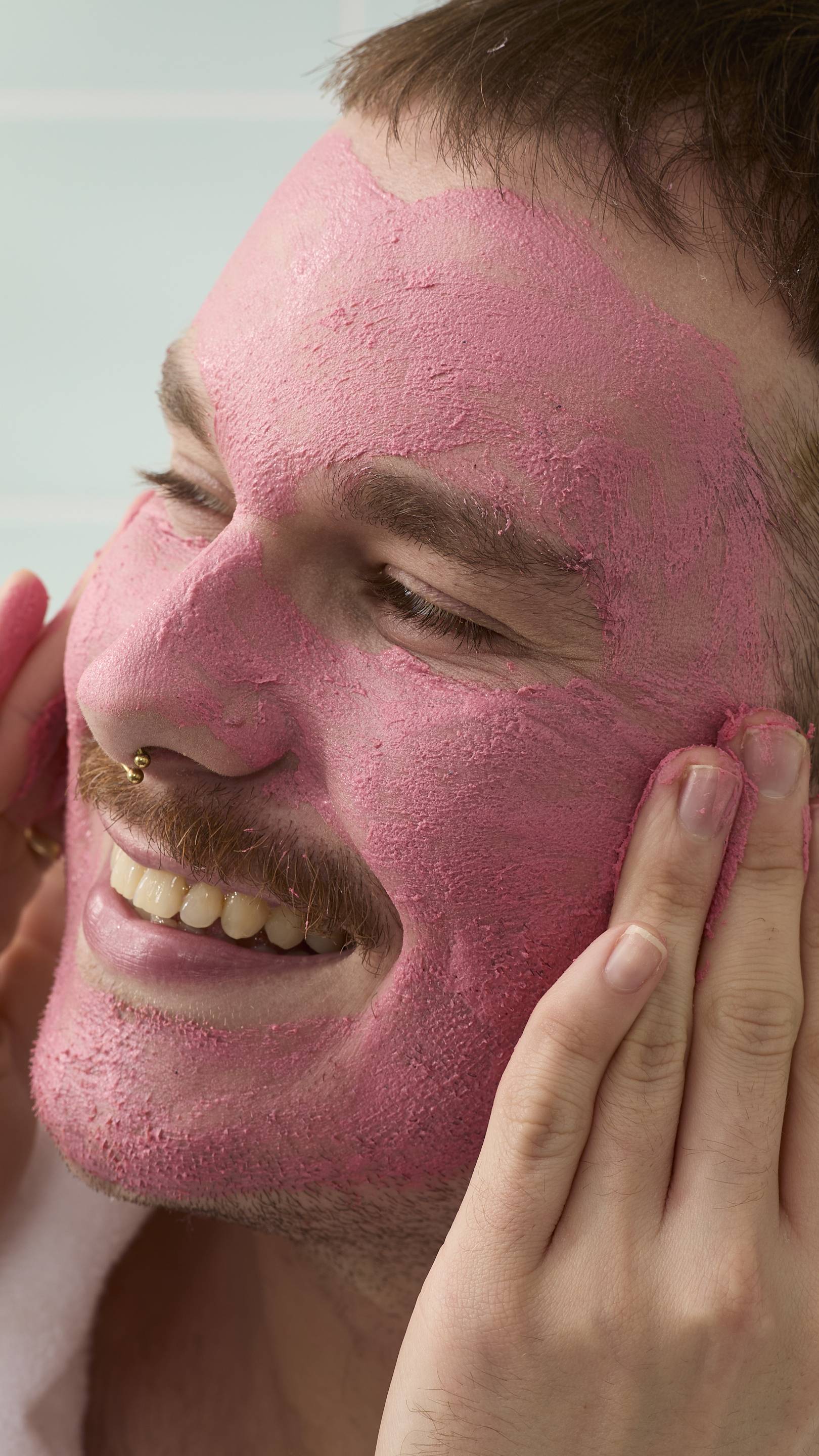 A close-up, profile view of a smiling model with a moustache and facial piercings. Their face is almost entirely covered in a thick, textured pink "Ladybird" face cleanser, which they are massaging into their cheek with their fingertips.