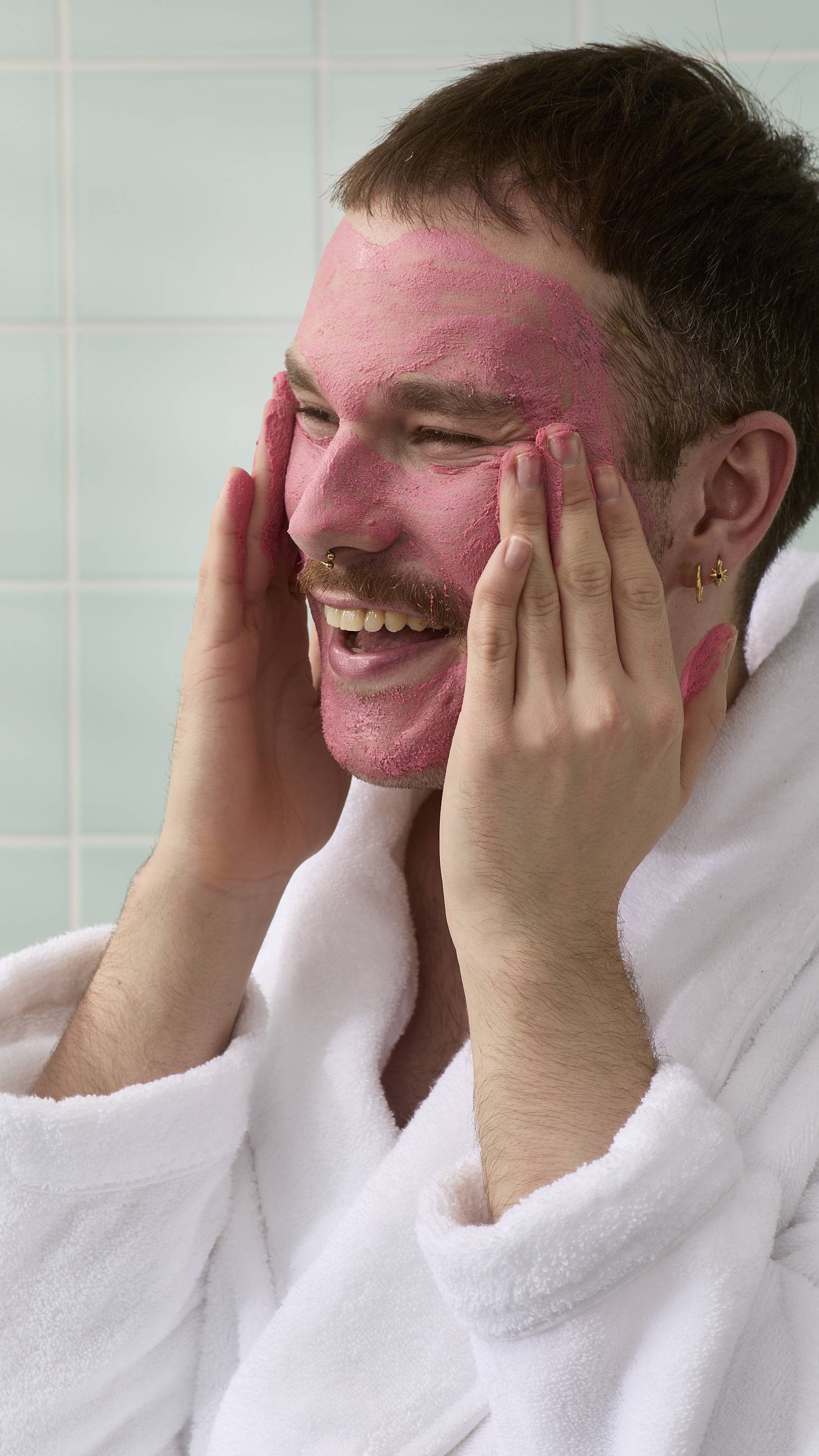 A profile view of a smiling model with a moustache and facial piercings. Their face is almost entirely covered in a thick, textured pink "Ladybird" face cleanser, which they are massaging into their cheek with their fingertips.