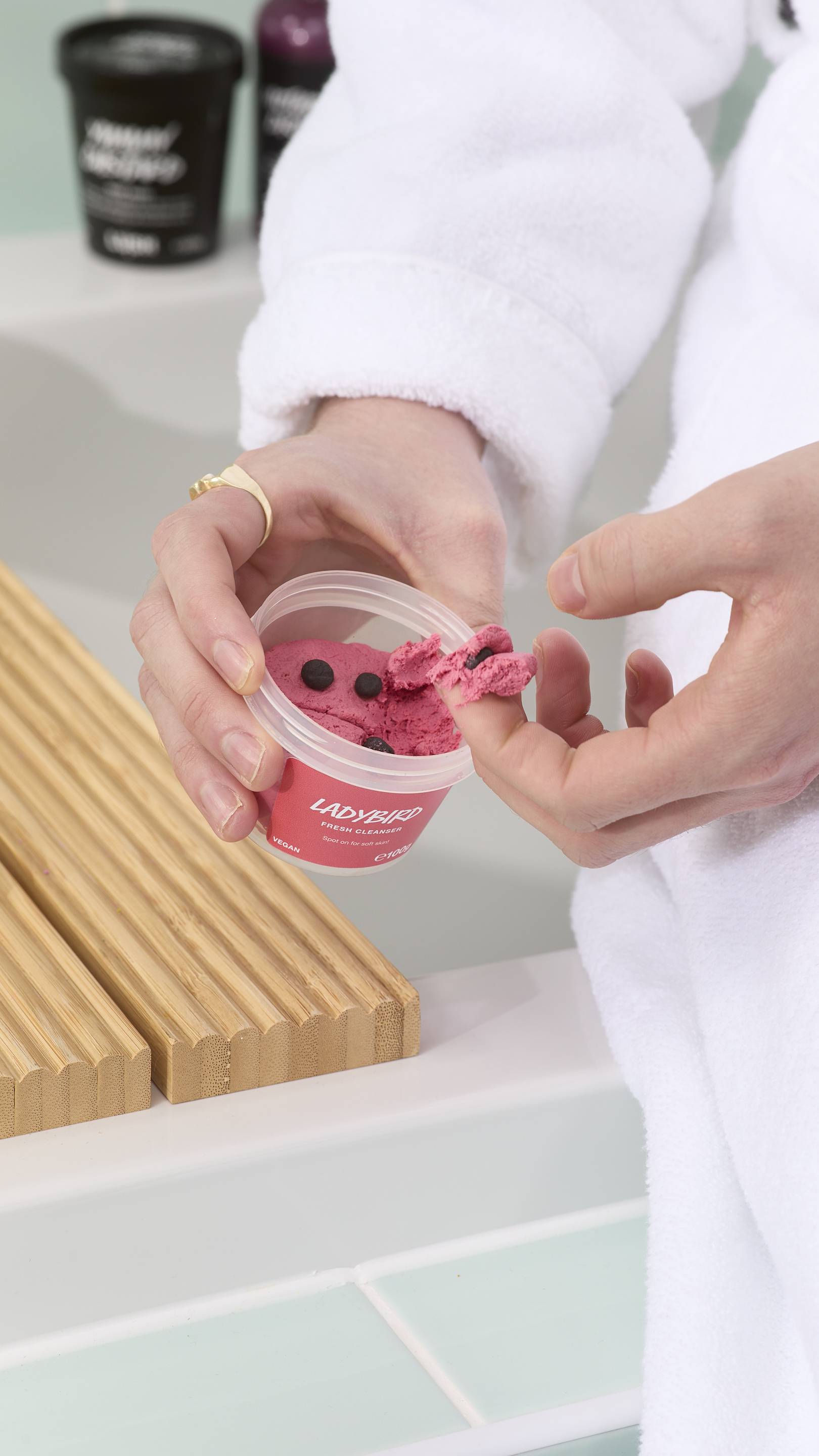 A close-up of a model in a white dressing gown holding a tub of "Ladybird" fresh facial cleanser. They have scooped a small amount of the thick, deep pink product onto their fingertip, which includes one of the small, dark exfoliating spheres.